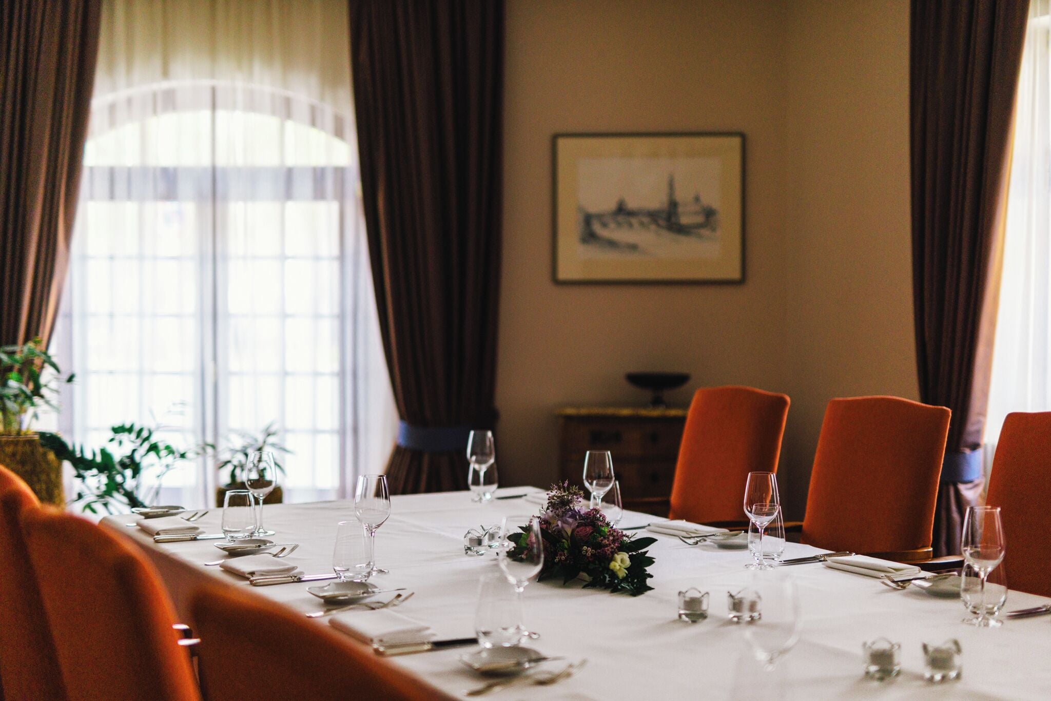 Mesa de comedor para seis personas, con mantel blanco, cubiertos y copas de vino, en una habitación con sillas naranjas y un cuadro en la pared.