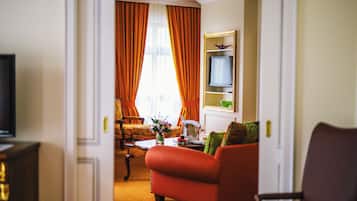 A hotel room with a sofa, a television on a stand, a dining table with a vase of flowers, and a large window with curtains.