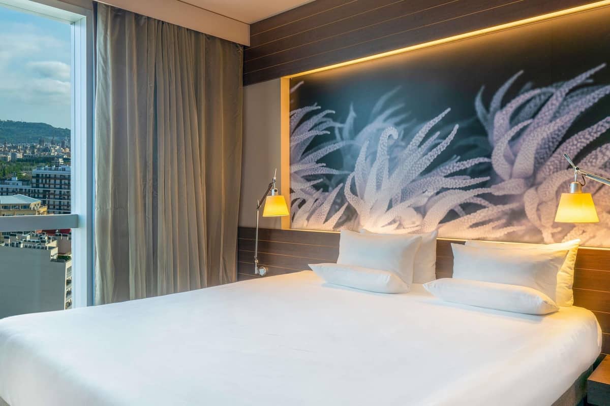 A hotel room with a large bed, two bedside lamps, a city view through a window, and a wall mural.