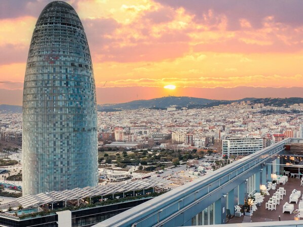 A modern cityscape with a prominent glass skyscraper, a bridge with outdoor seating, and a sunset in the background.