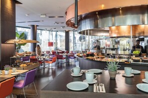 A modern restaurant with a buffet area, seating with purple and orange chairs, and a large kitchen in the background.