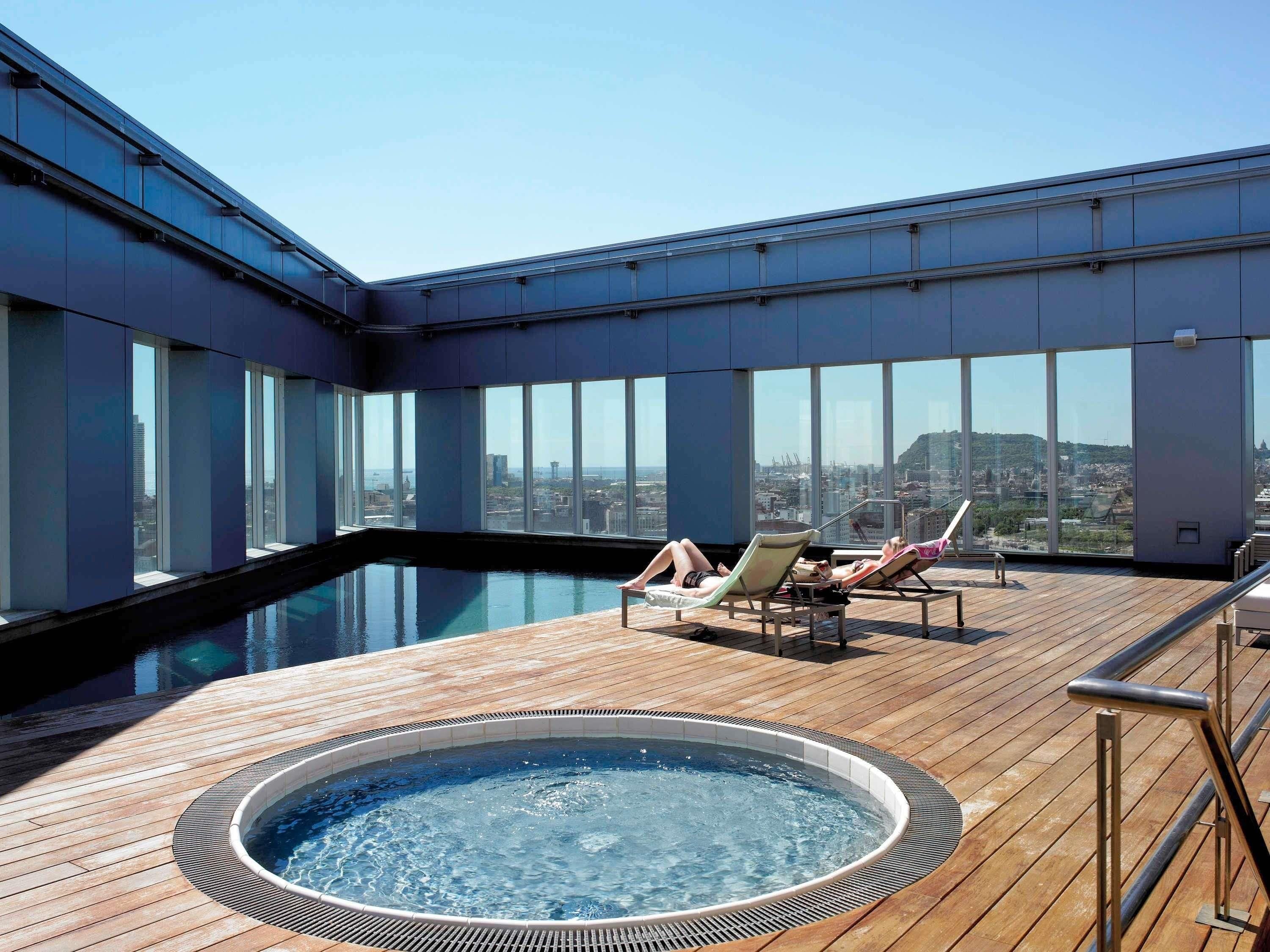 A rooftop pool area with a hot tub, lounge chairs, and a view of the cityscape.