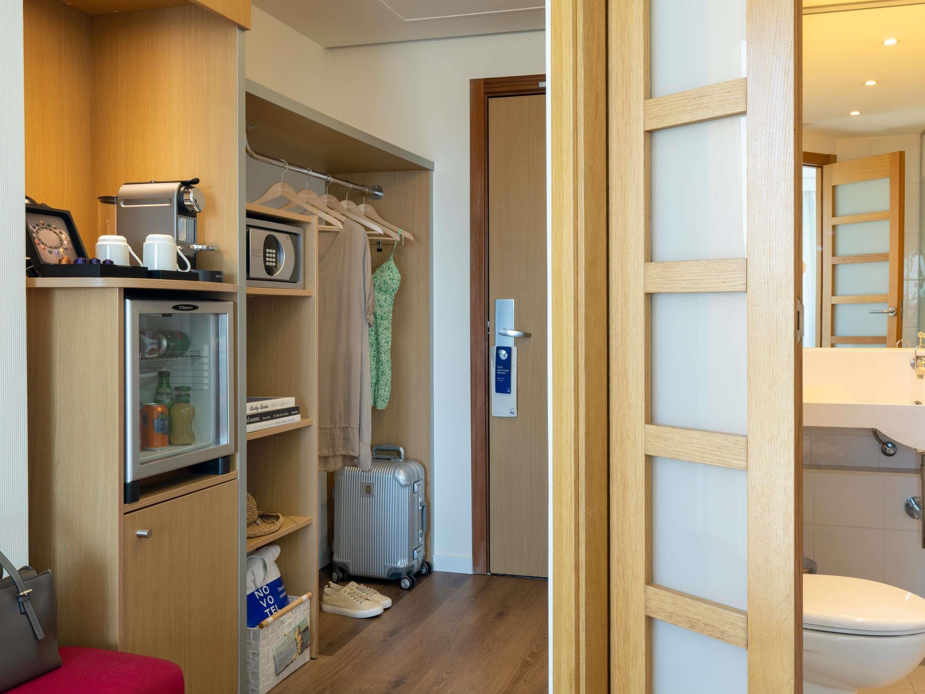 Une chambre d’hôtel compacte comprenant un coin cuisine, une armoire, une valise et une salle de bain.