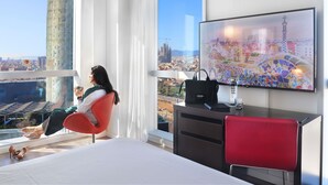 Moderni hotellihuone, jossa on suuri sänky, työpöytä, tuoli ja televisio, josta näkyy kaupunkimaisema.