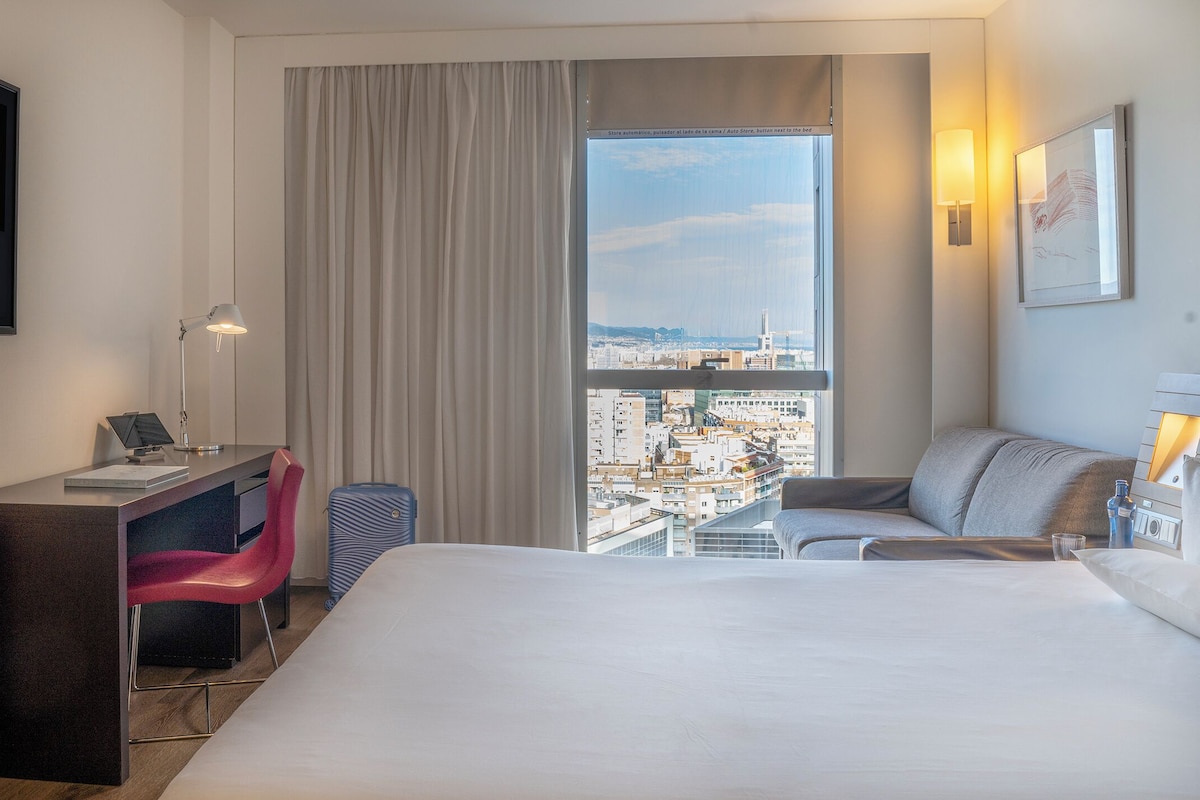 Ein Hotelzimmer mit Bett, Schreibtisch mit Stuhl, Fernseher und einem großen Fenster mit Blick auf die Stadt.