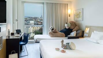 A hotel room with a large bed, a desk with a laptop, a teddy bear, and a city view.