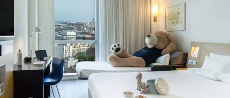 A hotel room with a large bed, a desk with a laptop, a teddy bear, and a city view.
