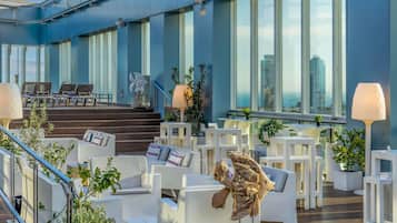 A modern outdoor seating area with white furniture, a staircase, and large windows offering views of the cityscape.