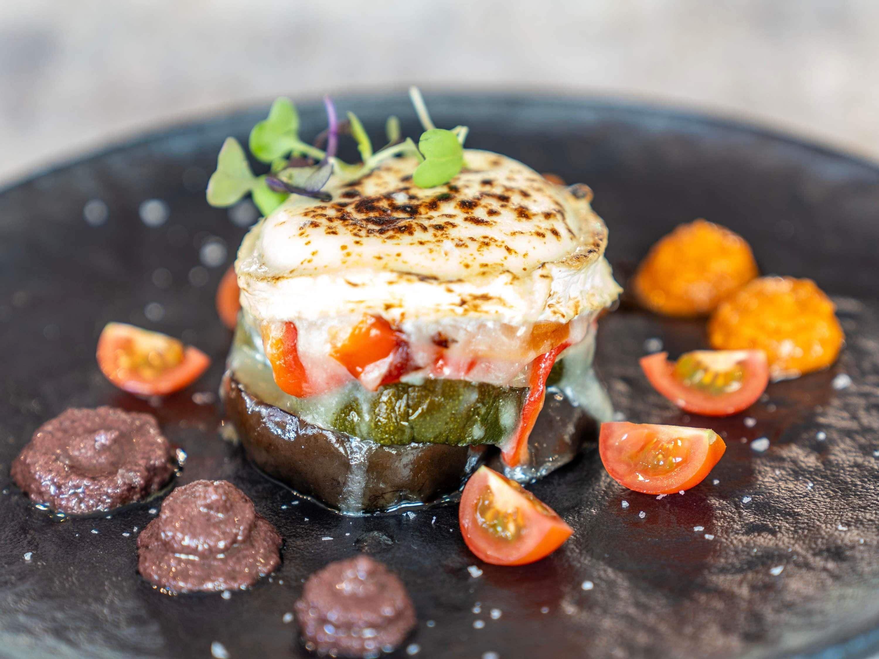 A dish with a creamy top, surrounded by vegetables and garnishes on a dark plate.