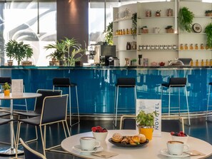 A modern hotel lobby with a blue reception desk, seating area with chairs, and a coffee station with pastries and cups.