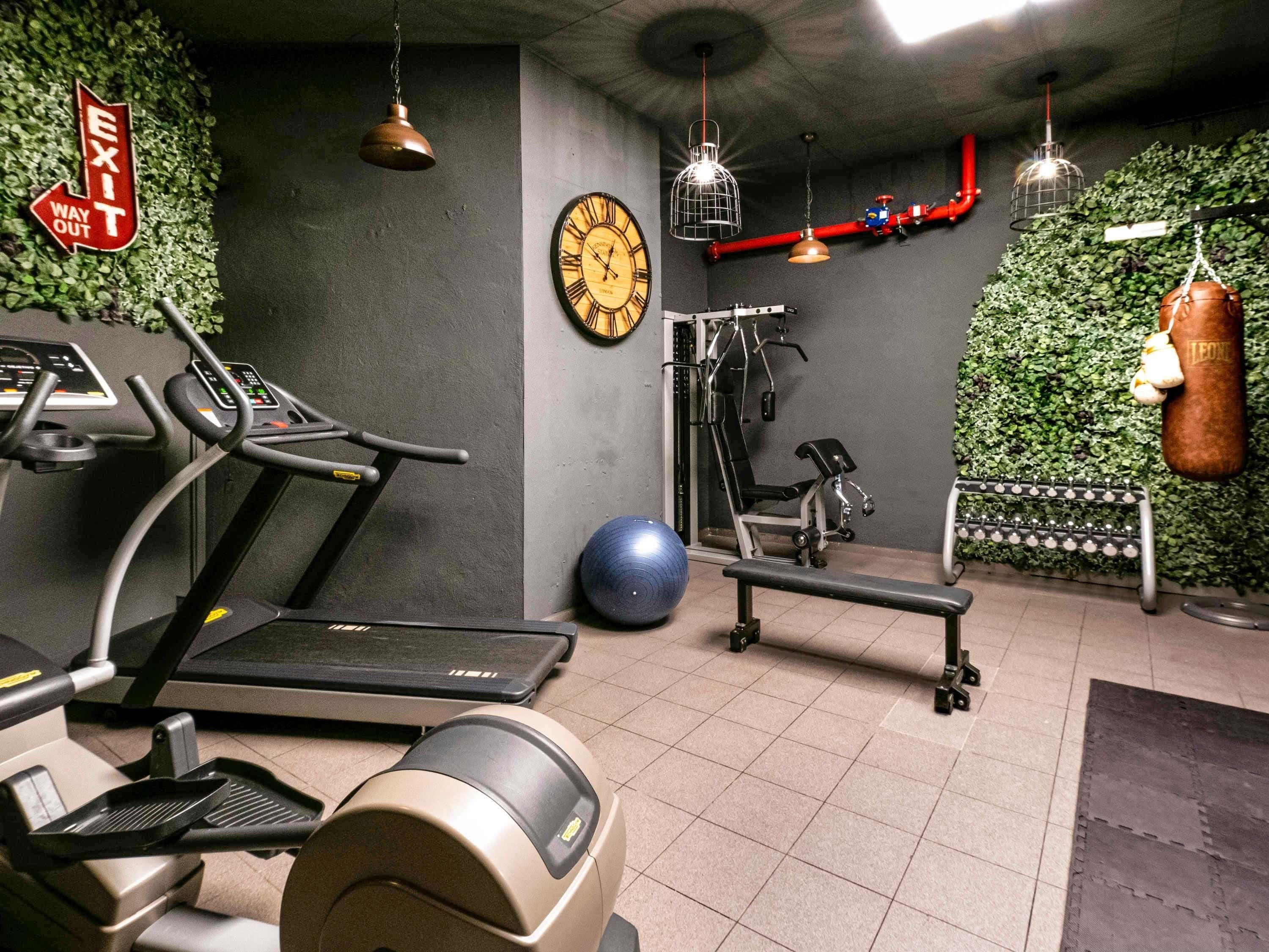 A gym with exercise equipment, a punching bag, a clock, and a wall with green foliage.