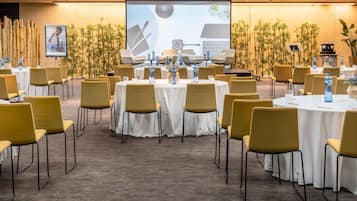 A conference room with round tables, yellow chairs, and a large screen displaying a presentation.