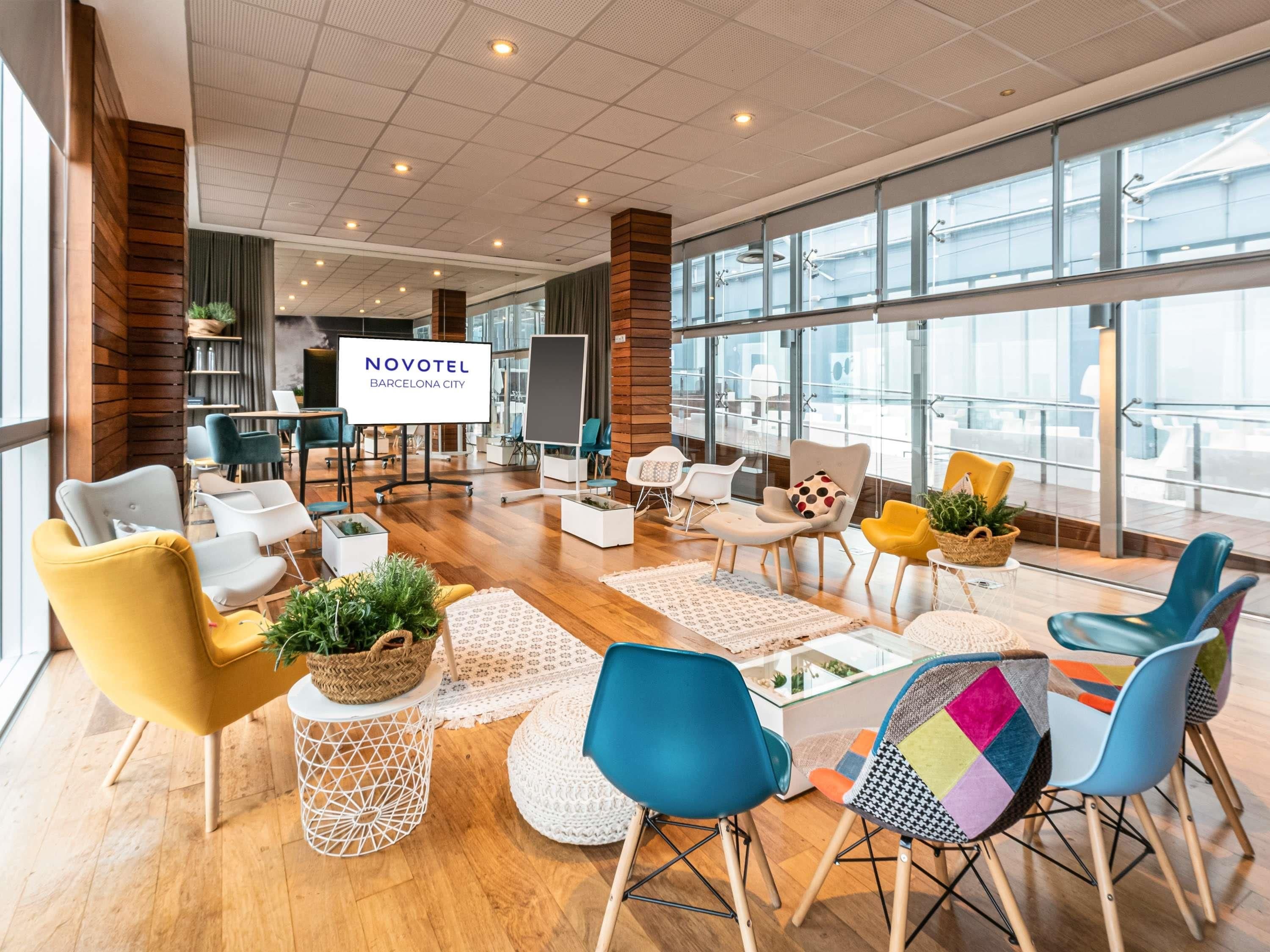 Un hall d’hôtel moderne, avec une grande enseigne indiquant « NOVOTEL BARCELONE CENTRE », des espaces de détente confortables et de grandes fenêtres offrant une vue sur la ville.