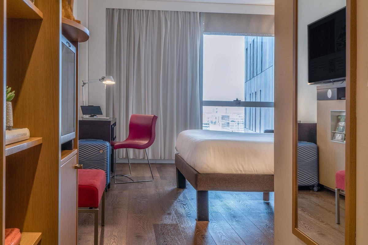 A hotel room with a bed, a desk, a chair, a television, and a window with a city view.