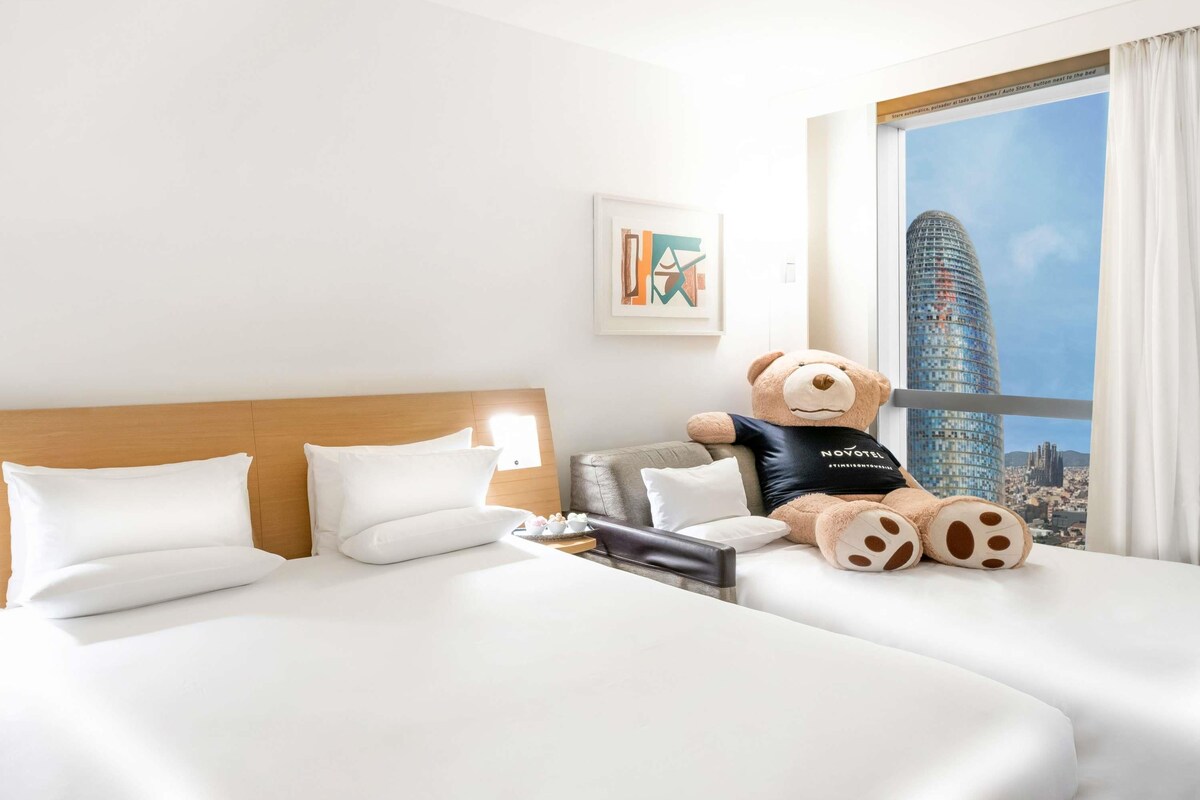 A hotel room with a bed, a teddy bear, a cityscape view, and a framed picture on the wall.