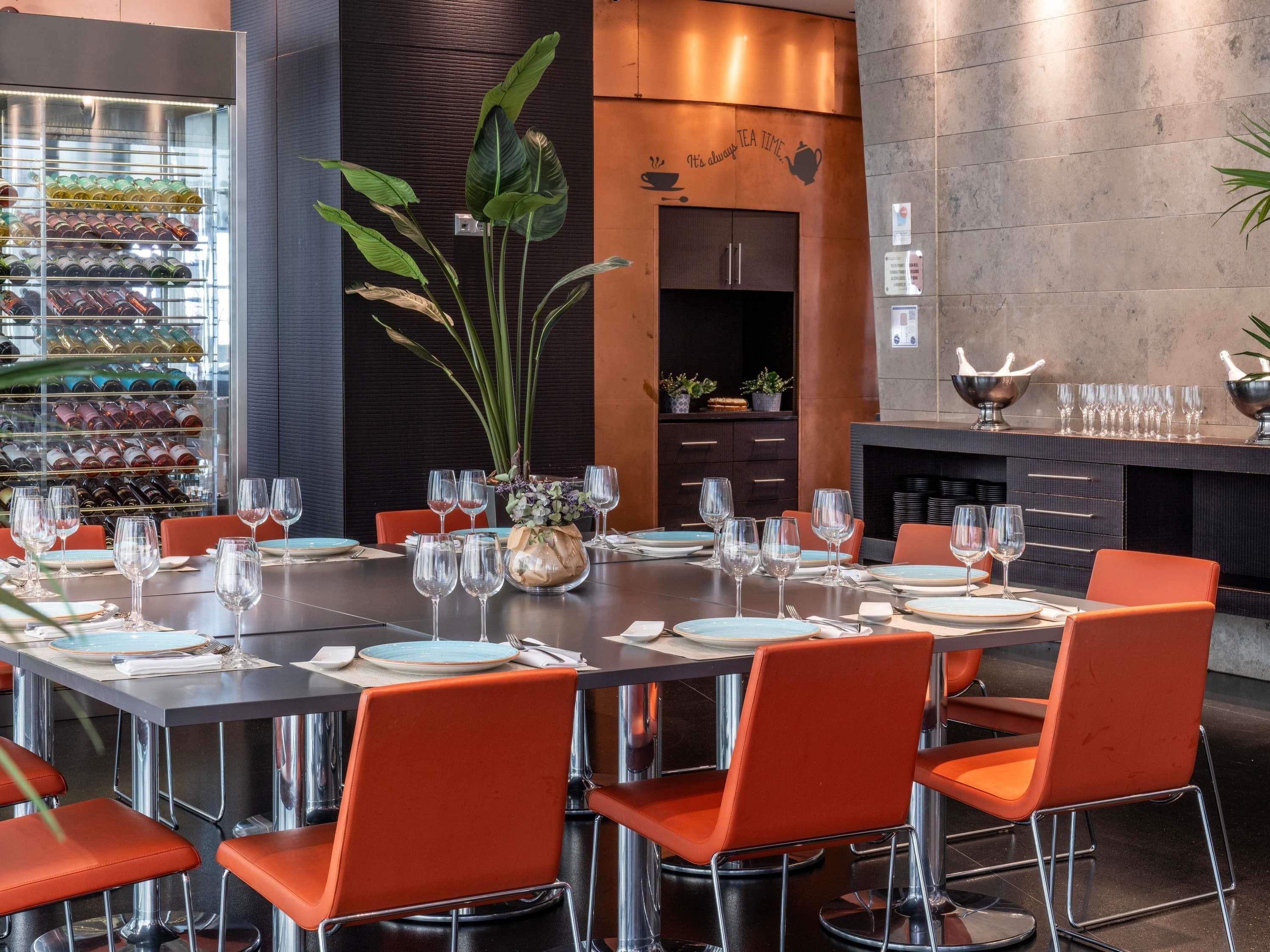 Un restaurante con una mesa redonda preparada para comer, rodeada de sillas naranjas, un enfriador de vino y una planta decorativa.