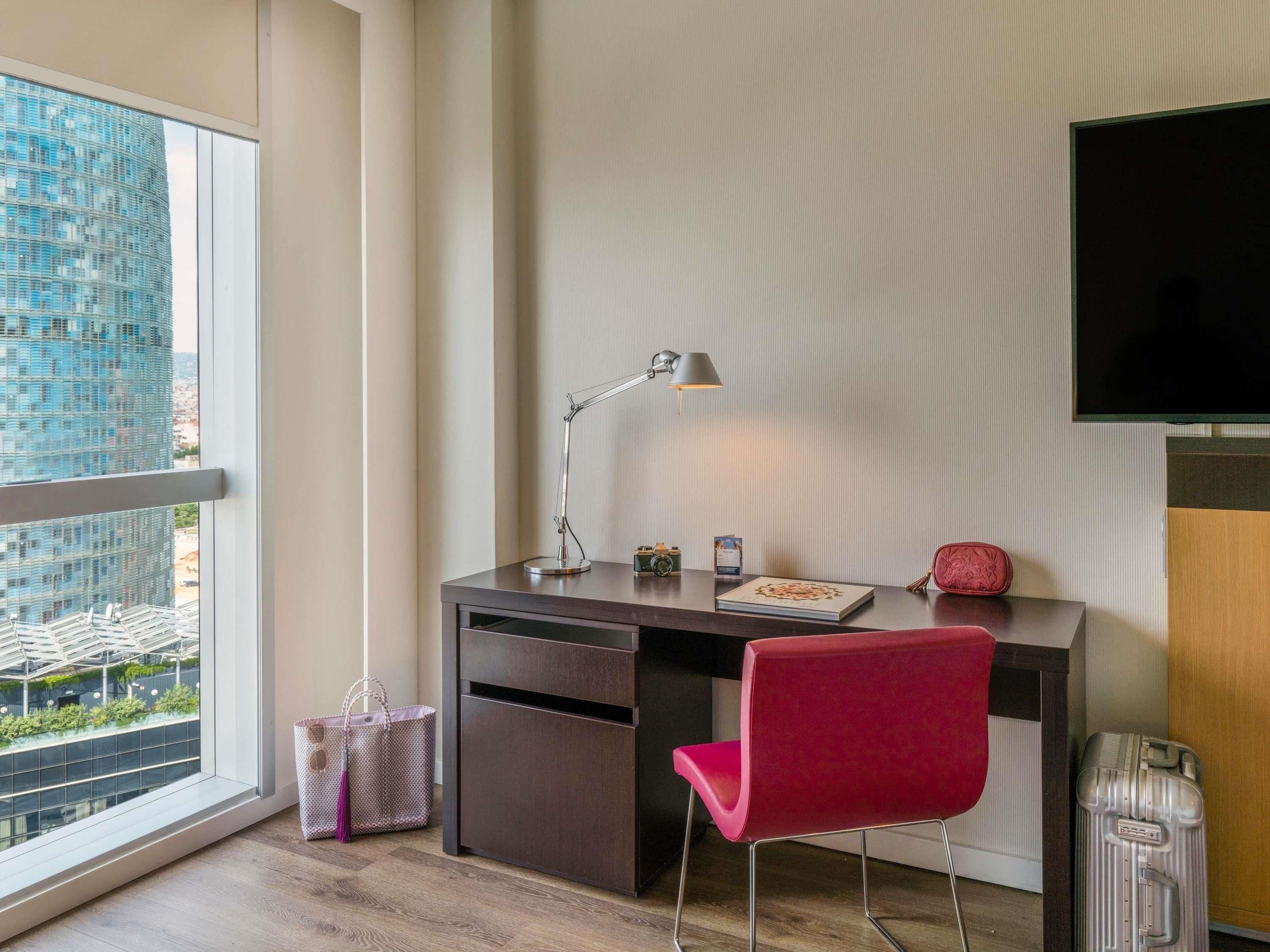 A modern hotel room with a desk, chair, and a large window offering a city view.