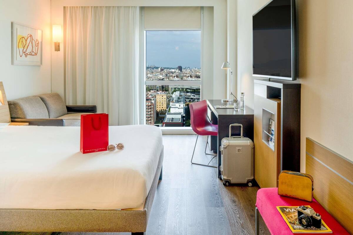 A hotel room with a bed, a desk, a chair, a TV, a suitcase, and a view of a cityscape.