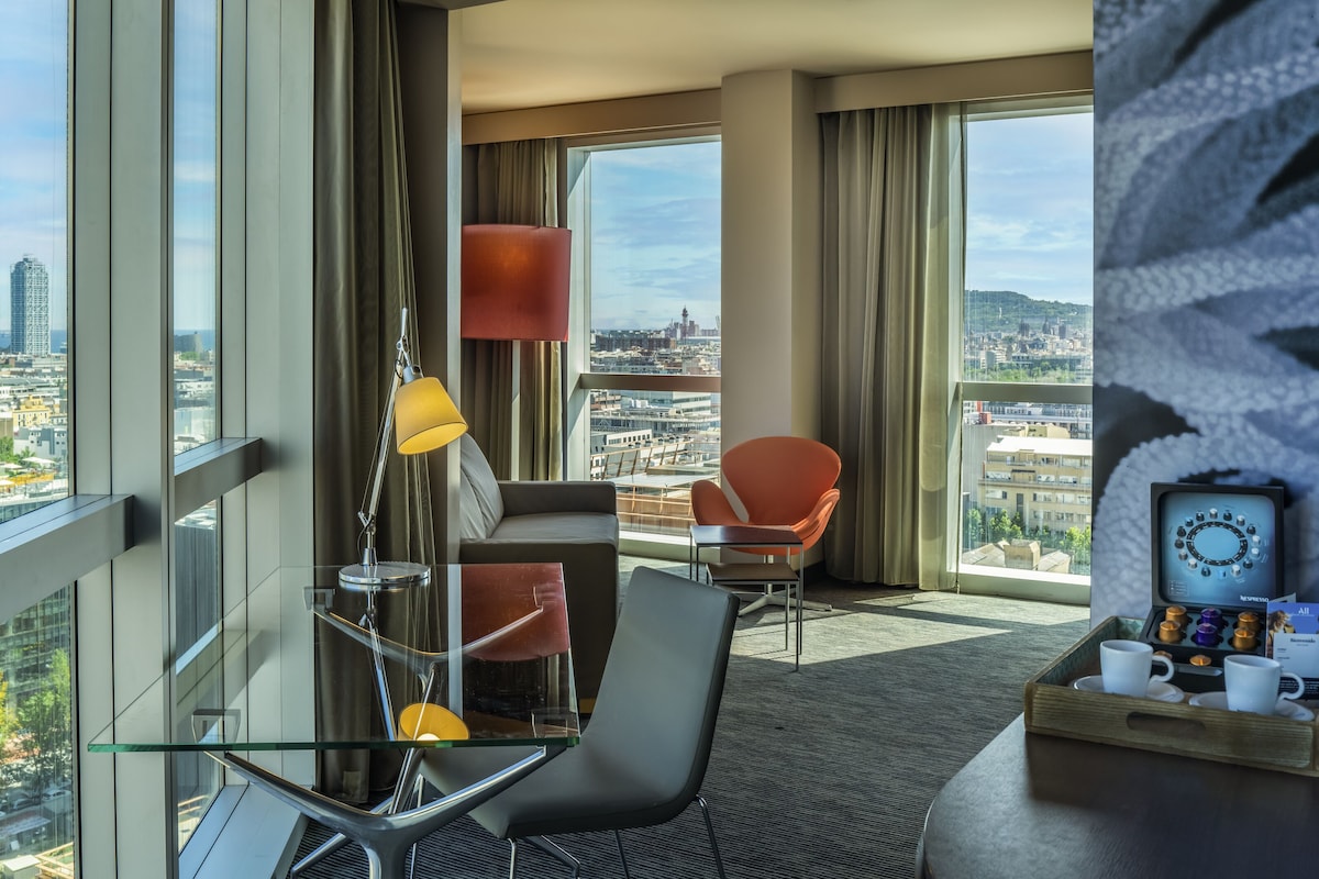 Ein modernes Hotelzimmer mit Blick auf die Stadt, einem Glastisch, einem roten Sessel, einer Lampe und einer Kaffeemaschine.