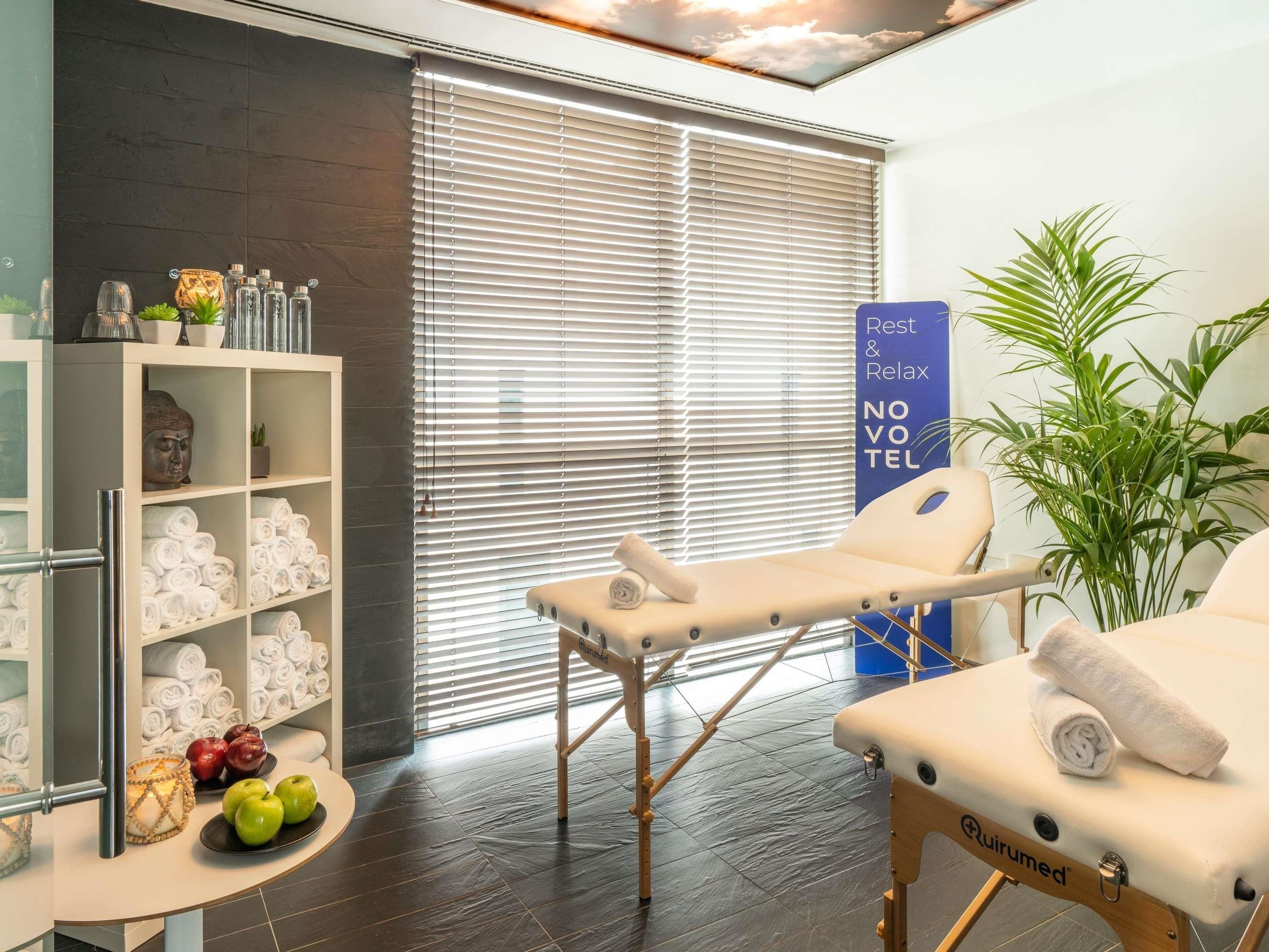 A modern spa room with a massage table, a shelf with towels and toiletries, and a large window with blinds.