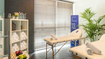 A modern spa room with a massage table, a shelf with towels and toiletries, and a large window with blinds.