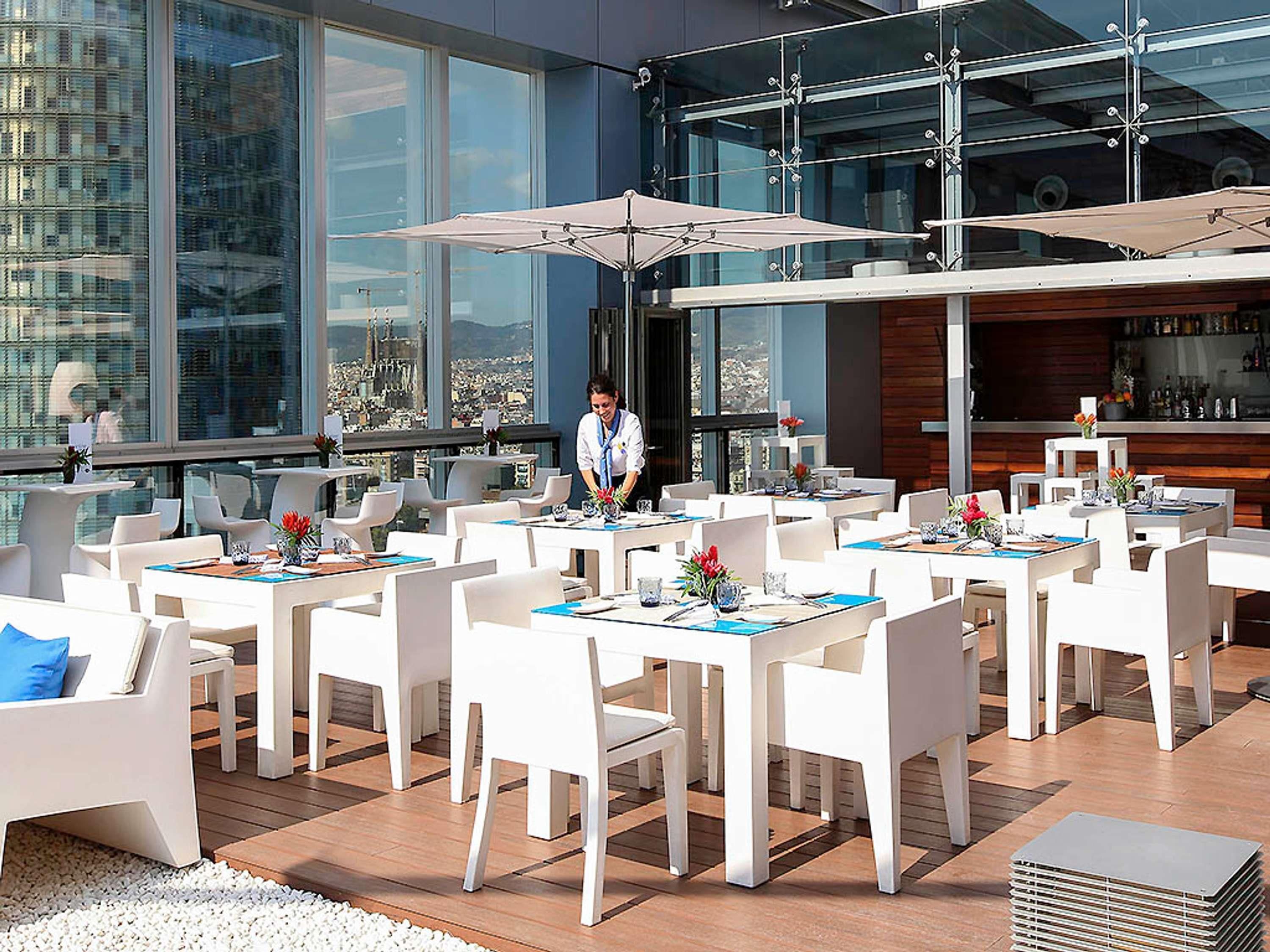 A modern outdoor dining area with white tables and chairs, large umbrellas, and a city view.