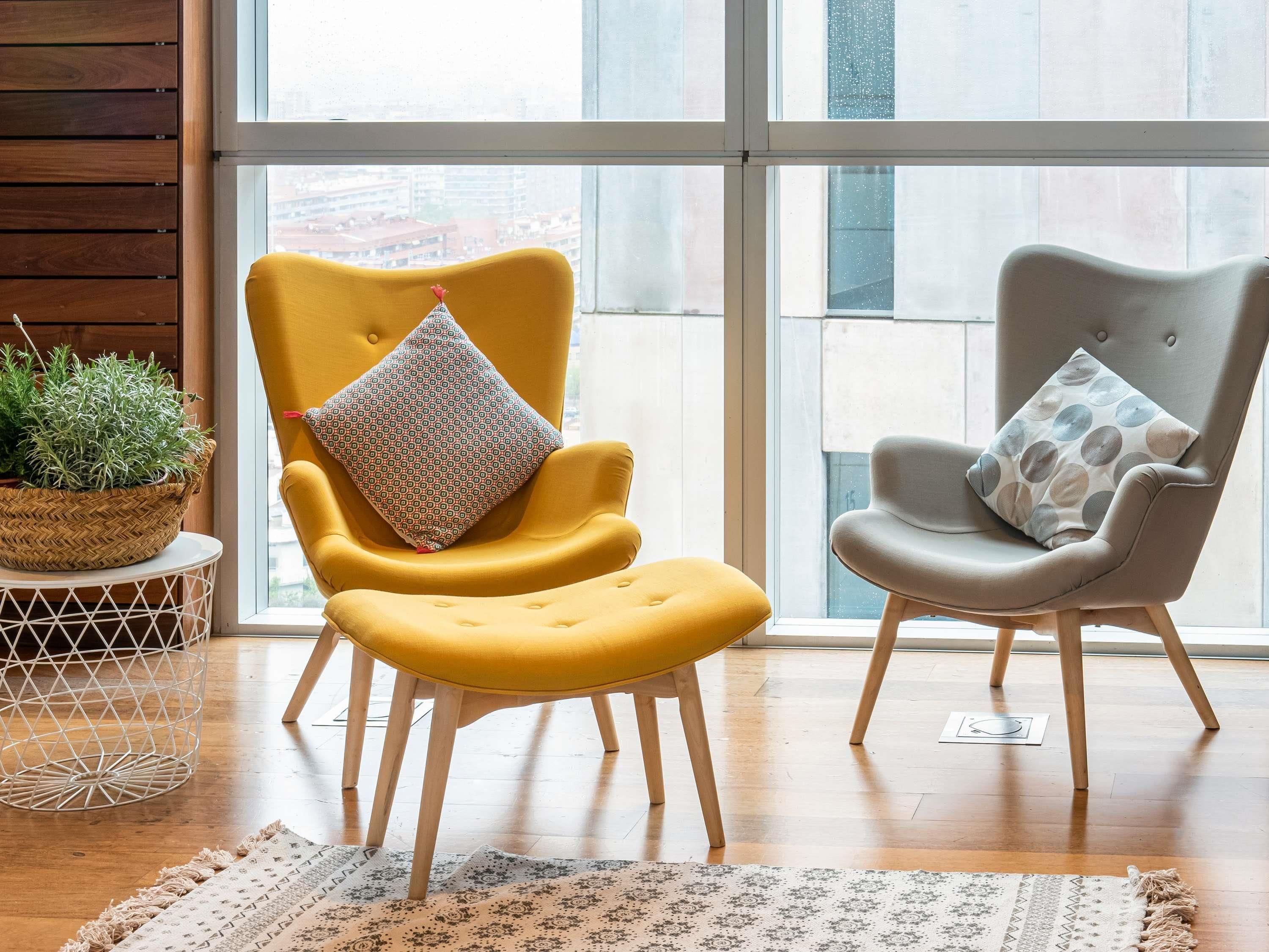Dos sillones con cojines estampados, una cesta tejida con follaje y una alfombra con diseño sobre un piso de madera.