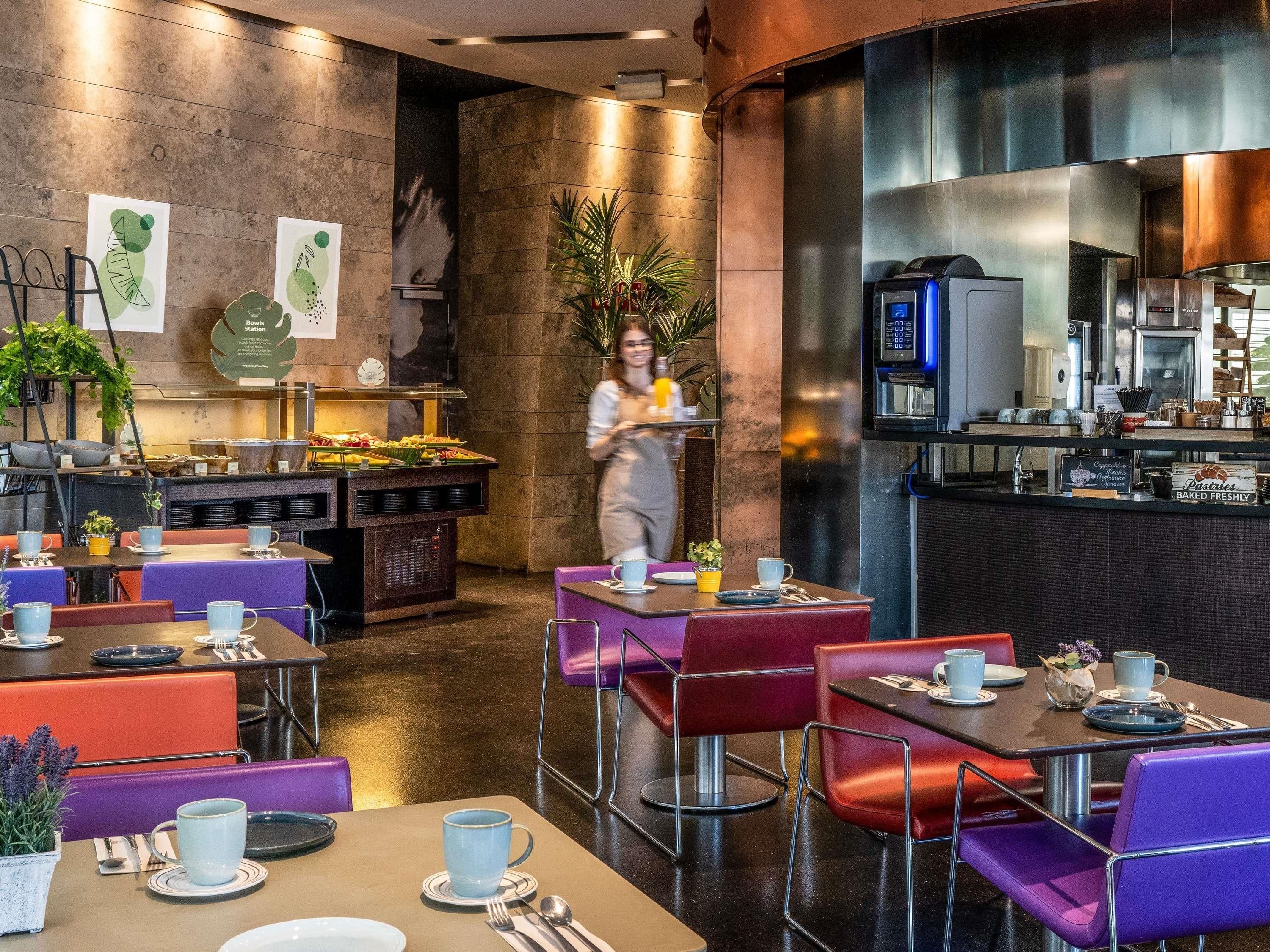 Un restaurante moderno con zona de buffet, mesas con sillas moradas y rojas, y una cocina a la vista.