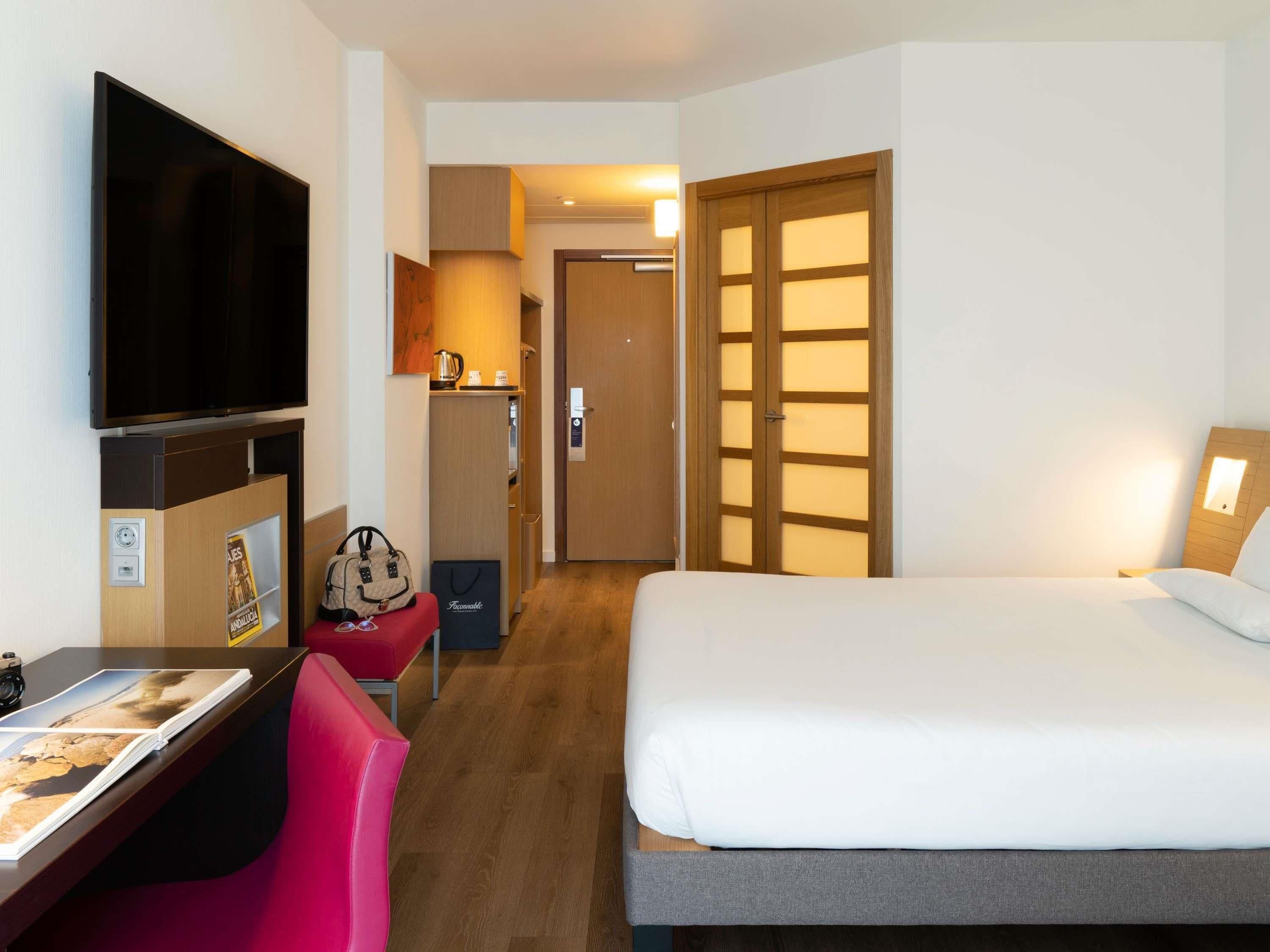 A hotel room with a bed, a desk with a chair, a television, a wardrobe, and a door leading to another room.