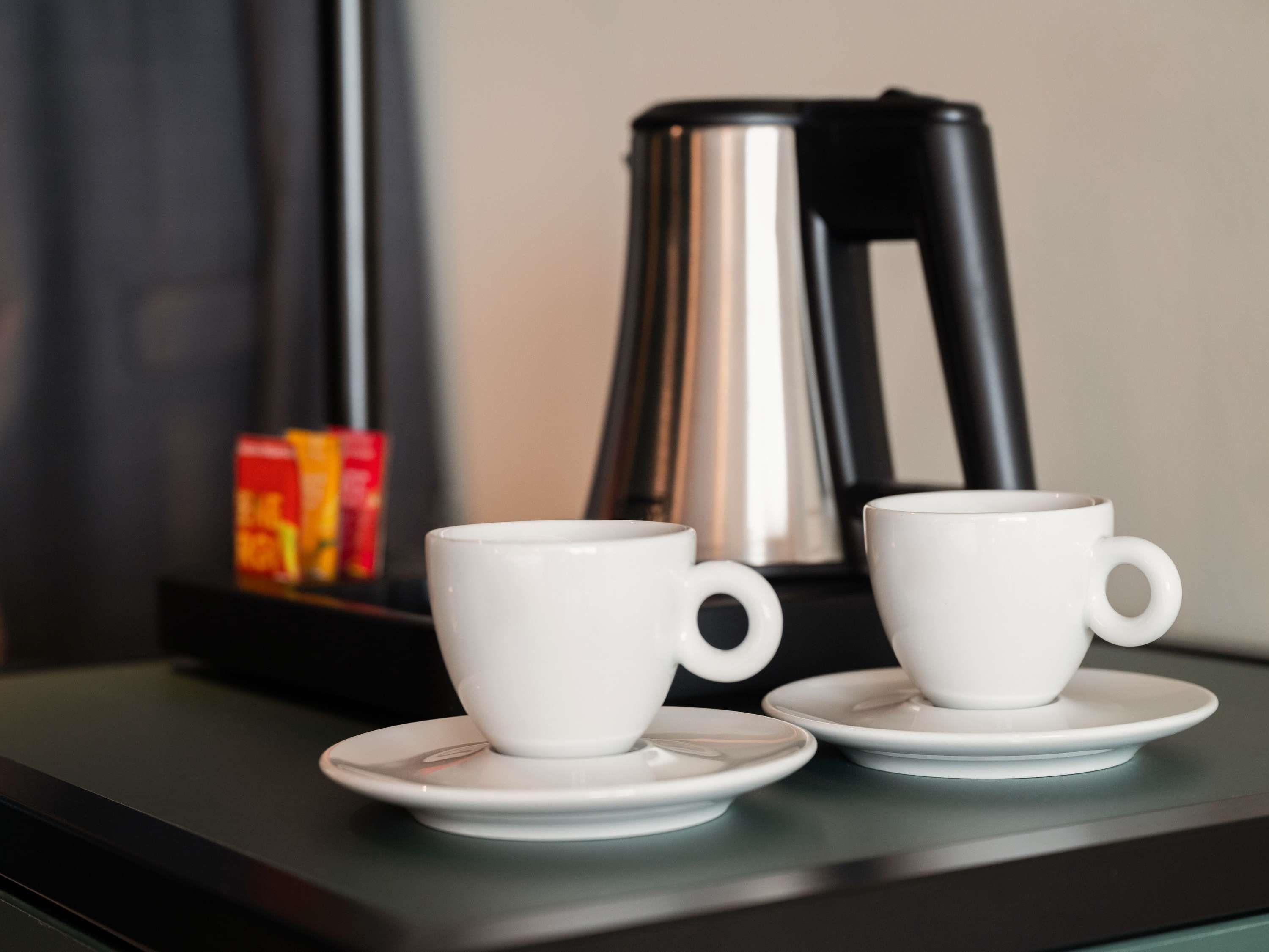 Zwei weiße Tassen auf Untertassen auf einer dunklen Oberfläche, im Hintergrund eine Kaffeemaschine.