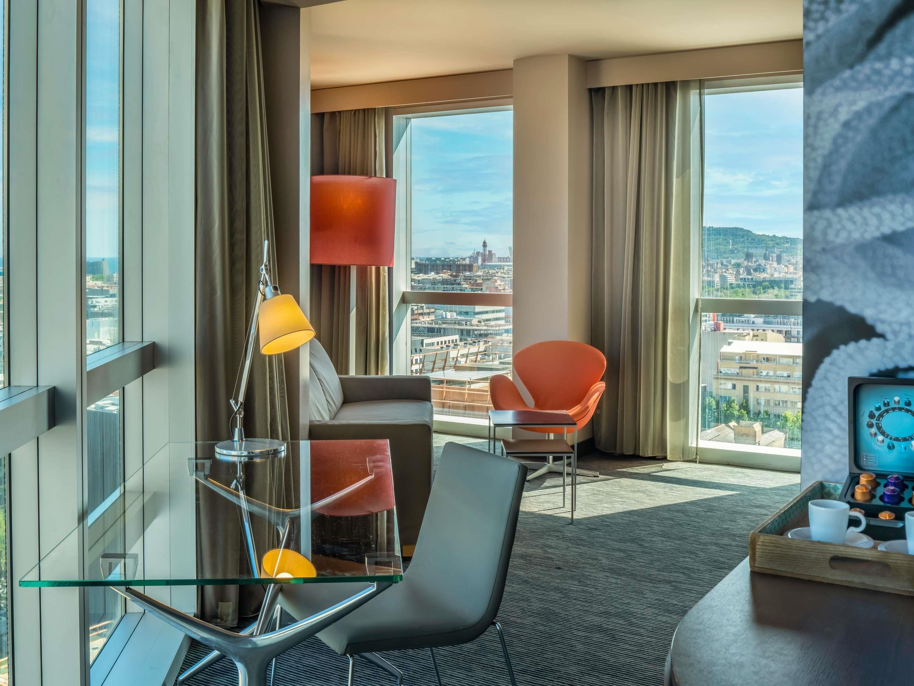 Habitación de hotel moderna con vista a la ciudad, una mesa de vidrio, un sofá, una silla y una bandeja con tazas.
