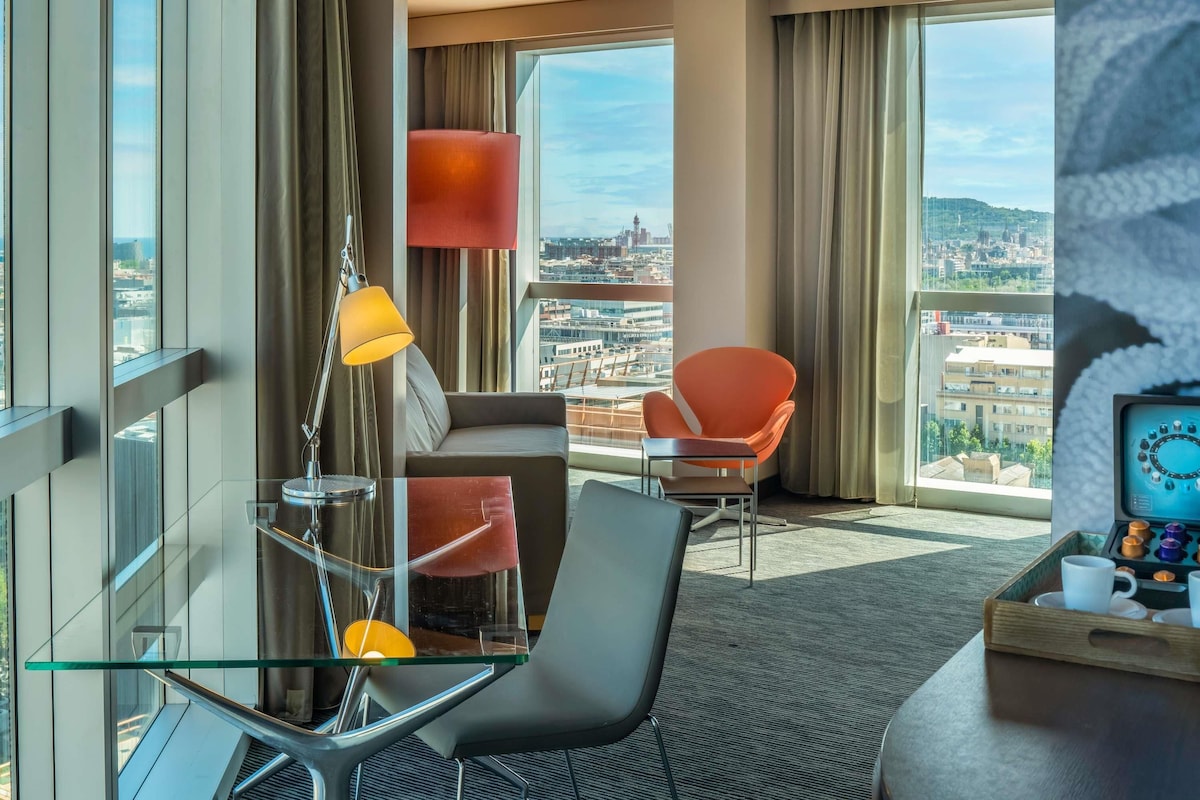 A modern hotel room with a city view, a glass table, a sofa, a chair, and a tray with mugs.