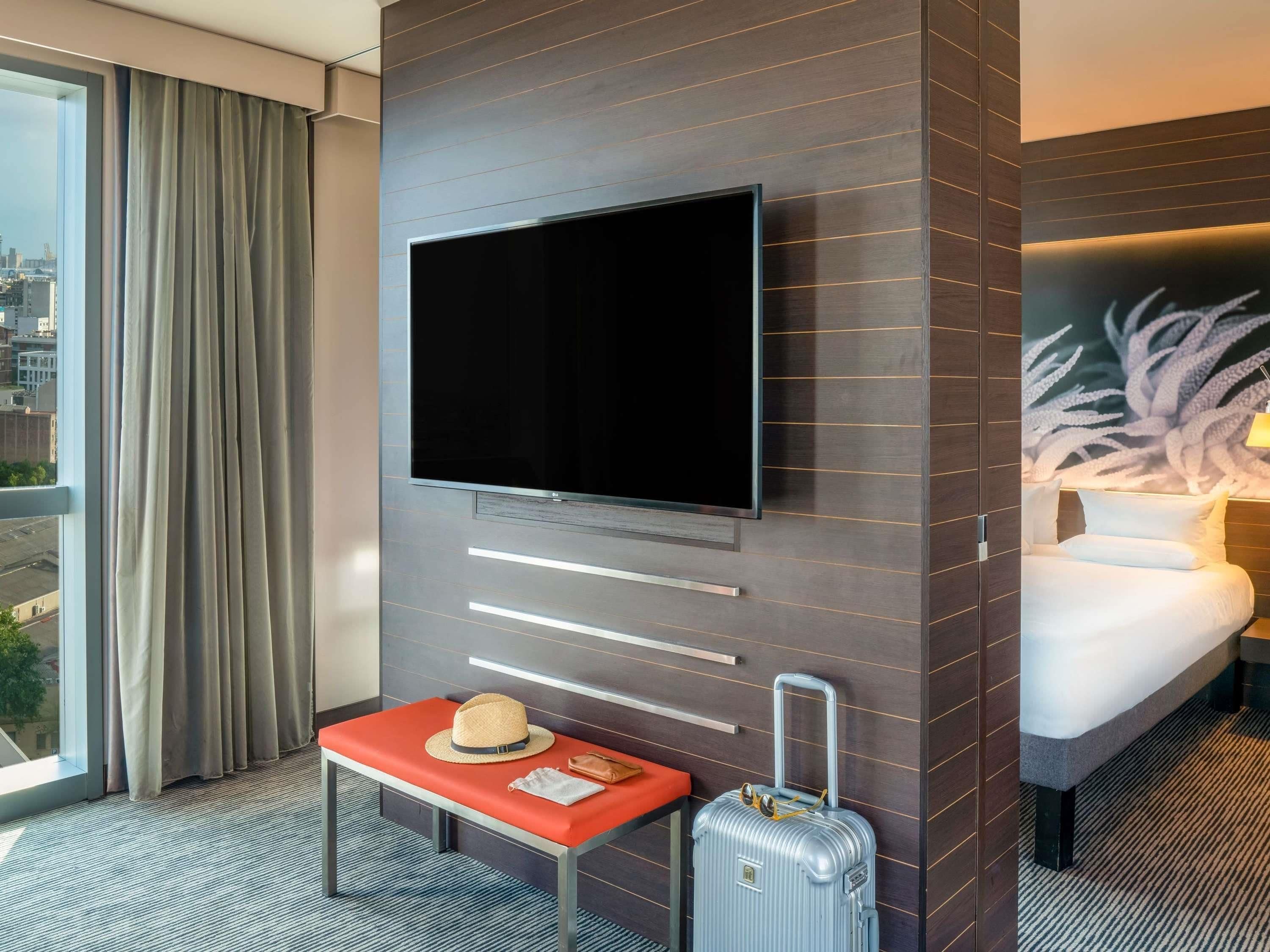 Une chambre d’hôtel équipée d’un téléviseur à écran plat fixé au mur, d’un lit avec une literie blanche, d’un banc sur lequel sont posés un chapeau et un sac, et d’une valise.