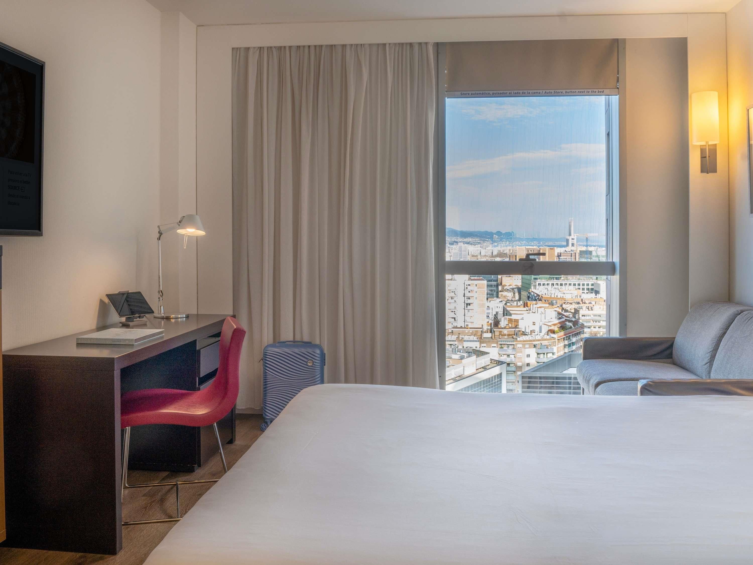 Ein Hotelzimmer mit Bett, Schreibtisch, Stuhl und einem großen Fenster mit Blick auf die Stadt.