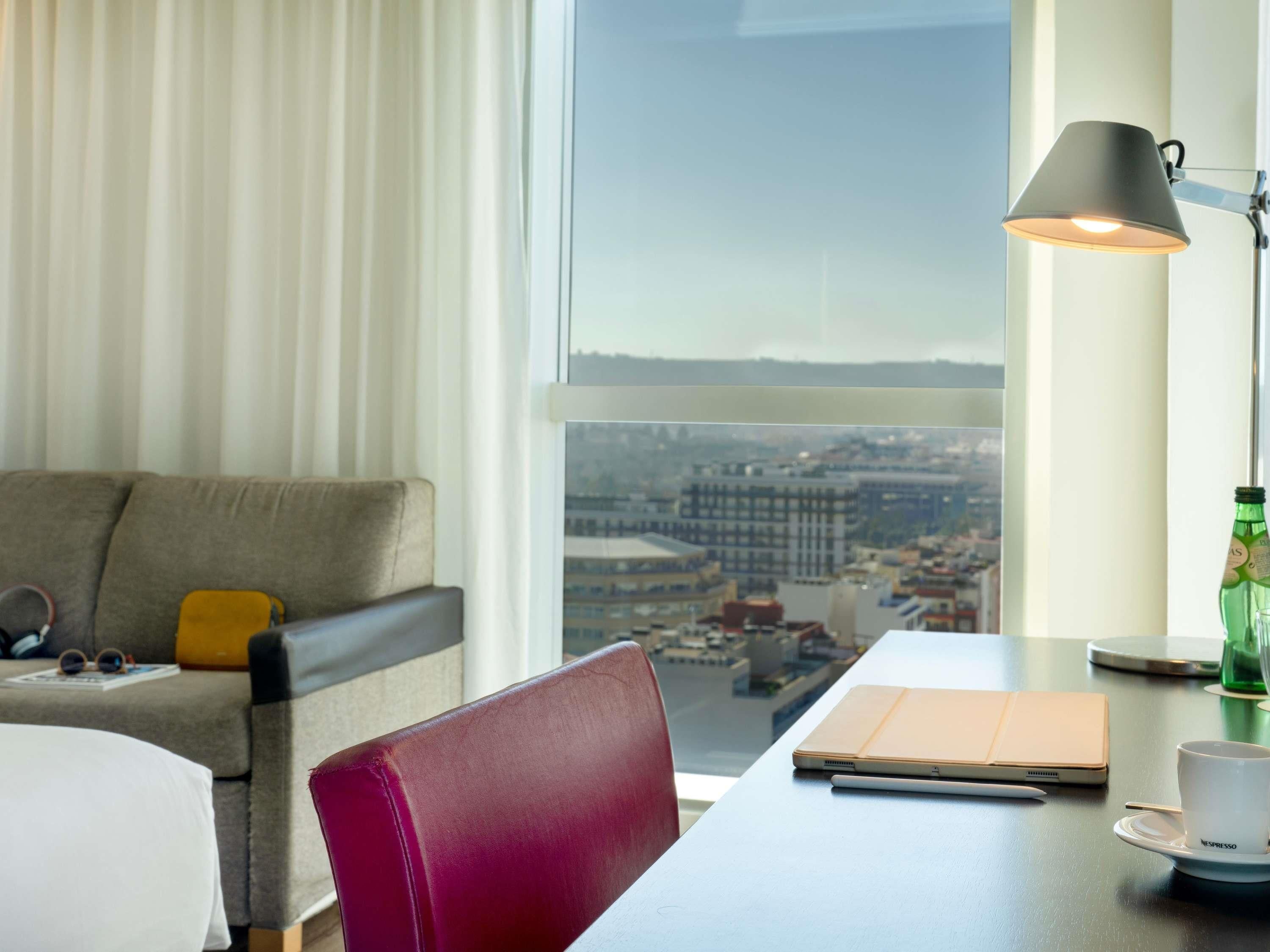 A hotel room with a bed, a desk with a laptop, a cup of coffee, and a view of a cityscape.