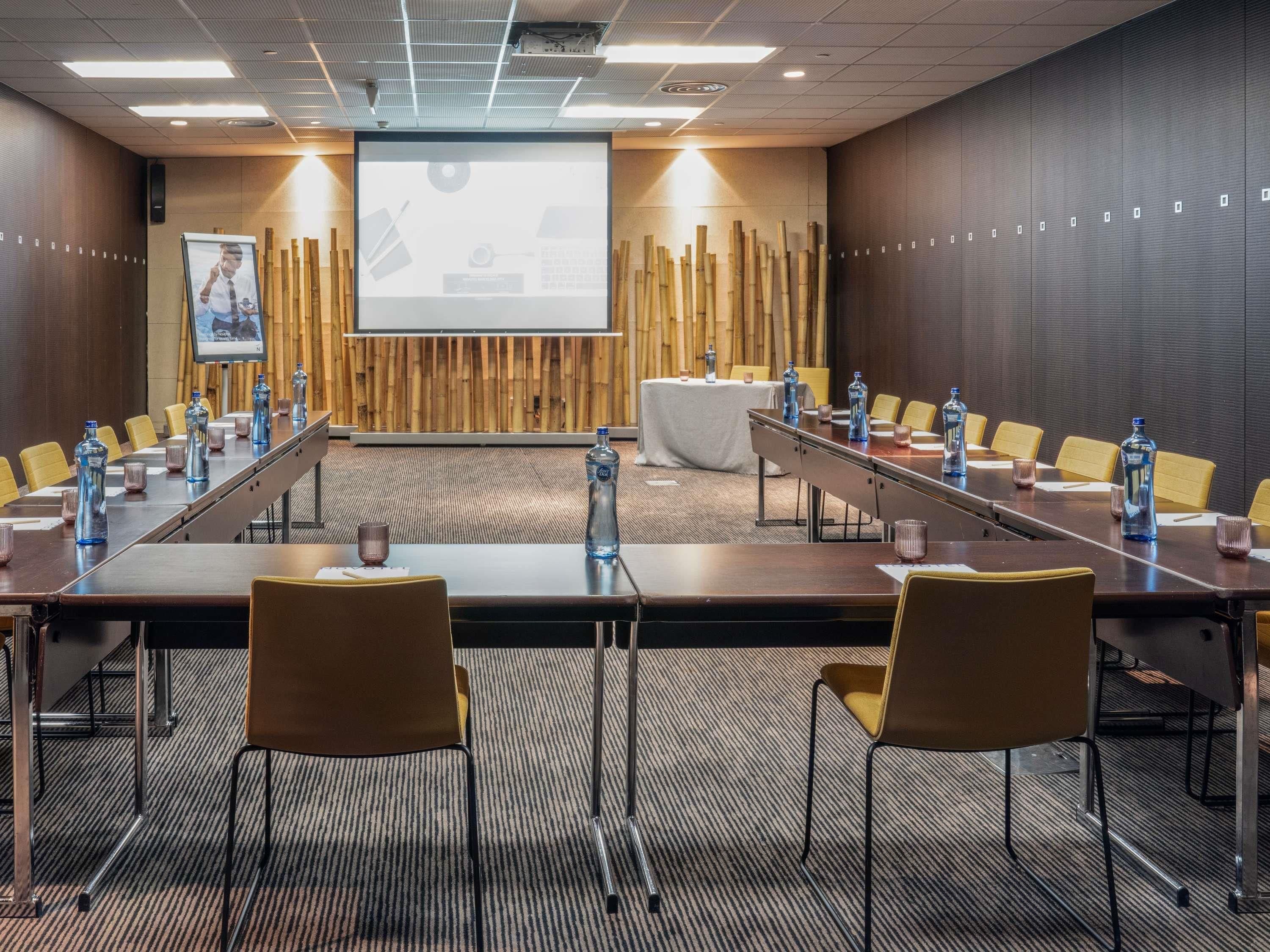Un salón de conferencias con una larga mesa, sillas, una pantalla de proyector y una pared de bambú.