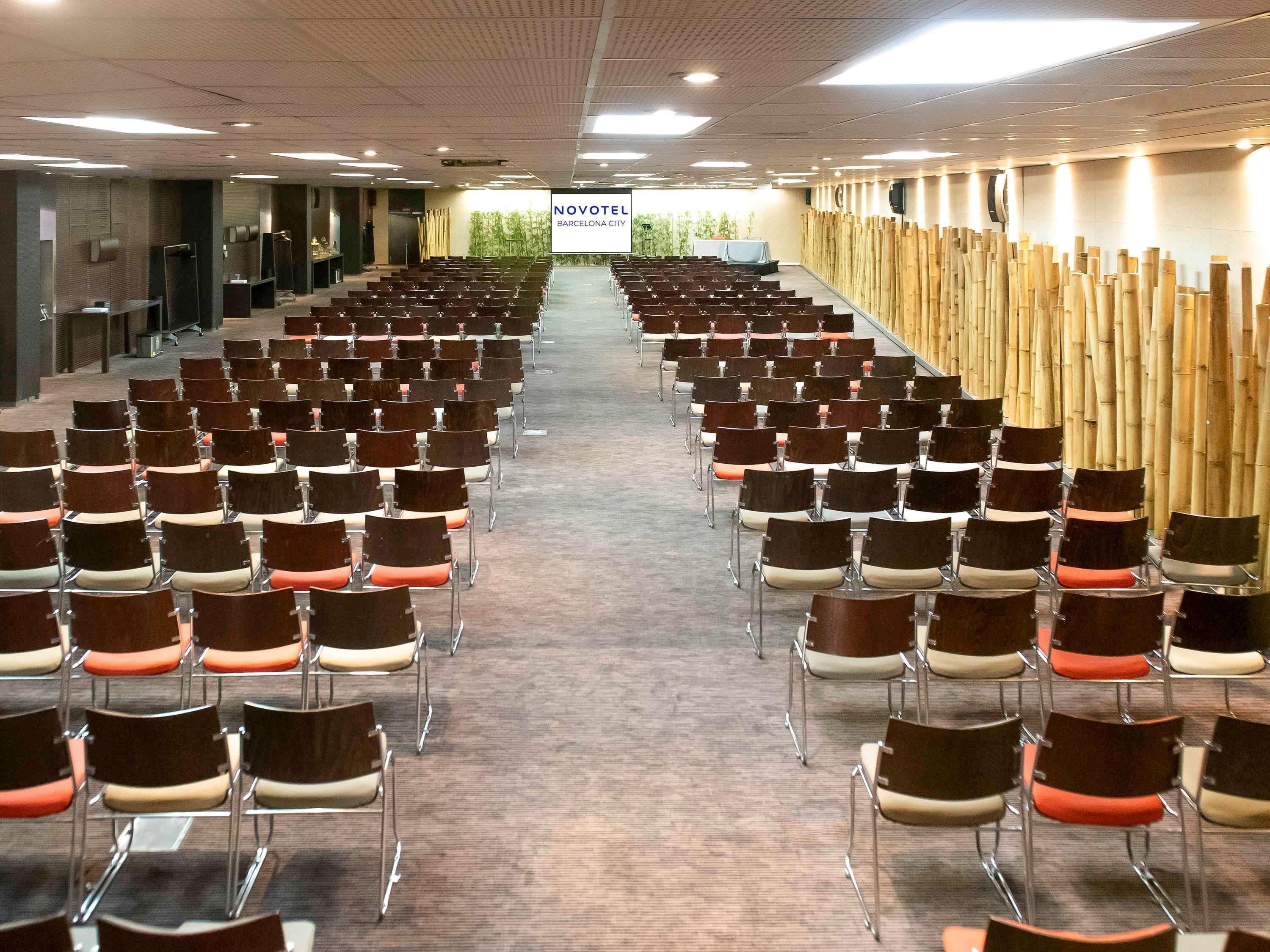 Un salón de conferencias con filas de sillas, un escenario y un cartel que dice 'NOVOTEL BARCELONA CITY'.