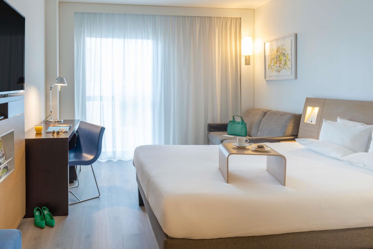 A hotel room with a bed, a desk with a chair, a television, and a window with curtains.