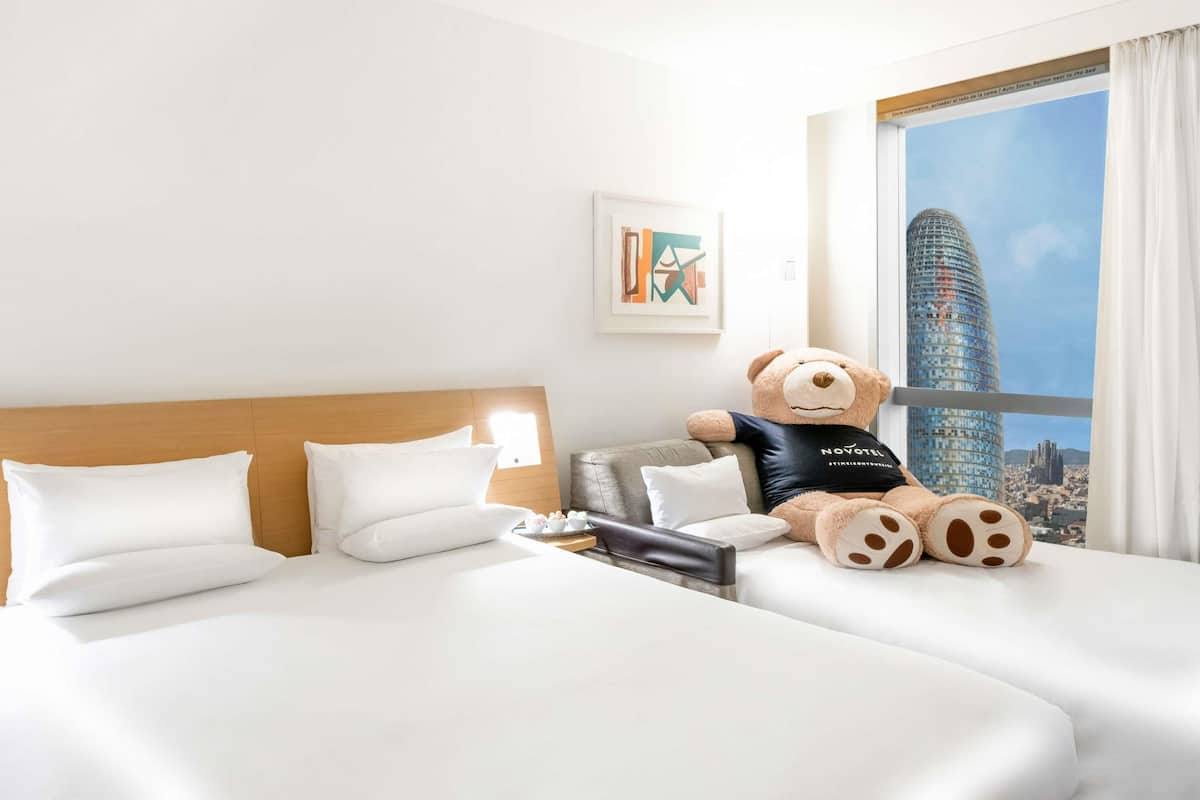A hotel room with a bed, a teddy bear, a cityscape view, and a framed picture on the wall.