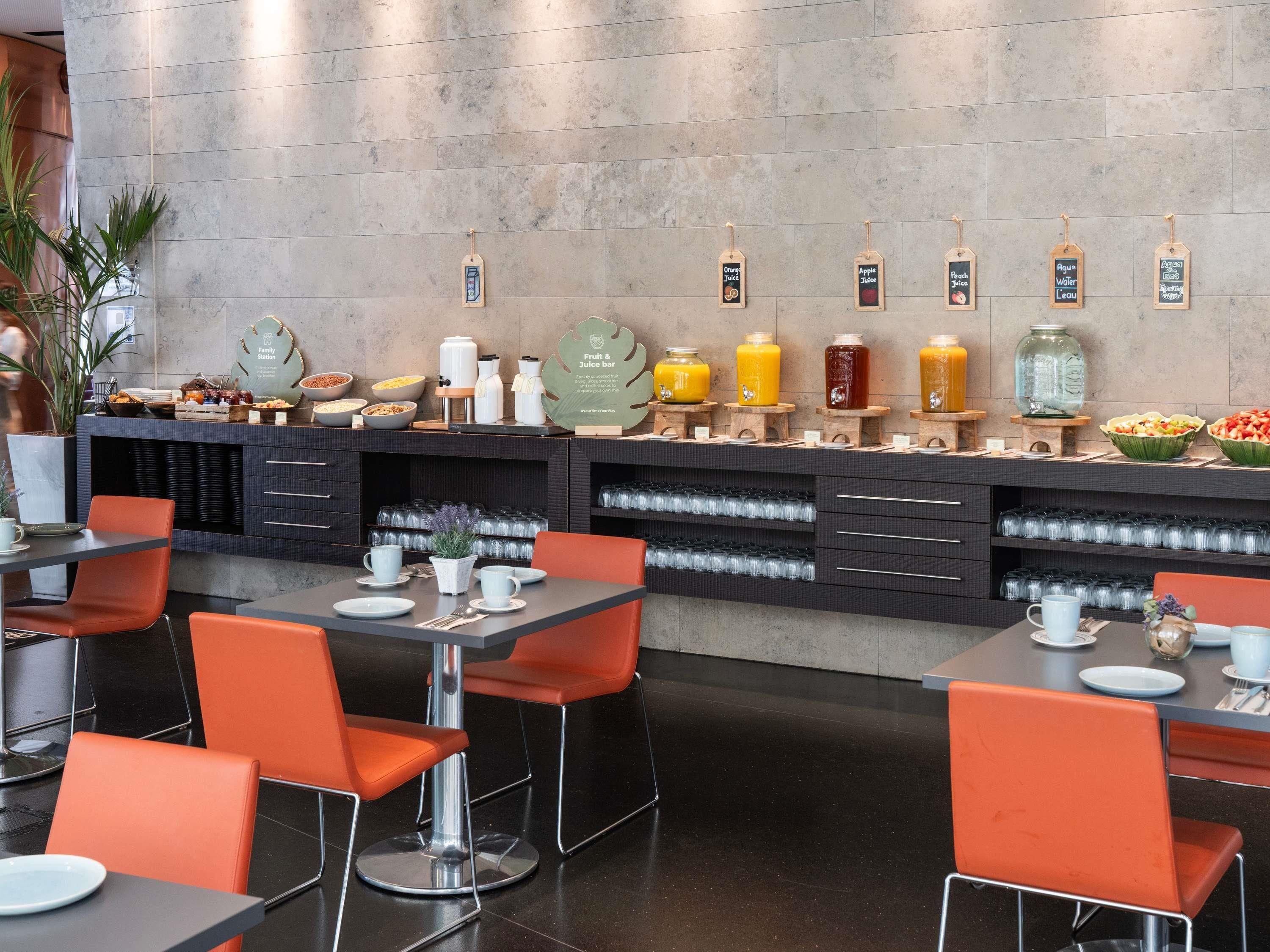 Een ontbijtbuffet met verschillende vruchtensappen en een koffiehoek. Er staan oranje stoelen en tafels met ontbijtartikelen.