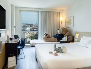 A hotel room with a large bed, a desk with a laptop, a teddy bear, and a city view.