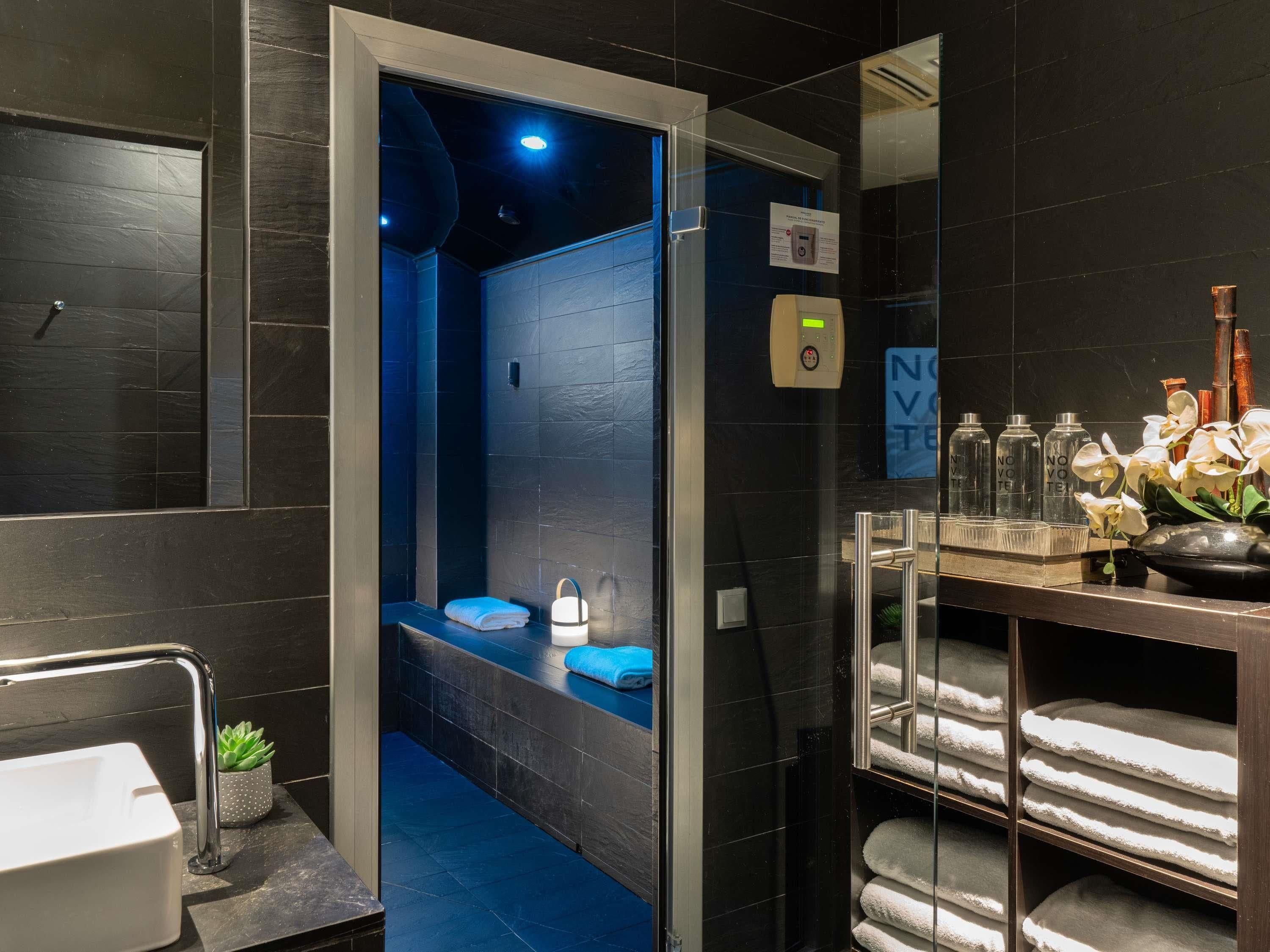 A modern bathroom with a blue-lit shower, a sink with a plant, and neatly arranged towels.