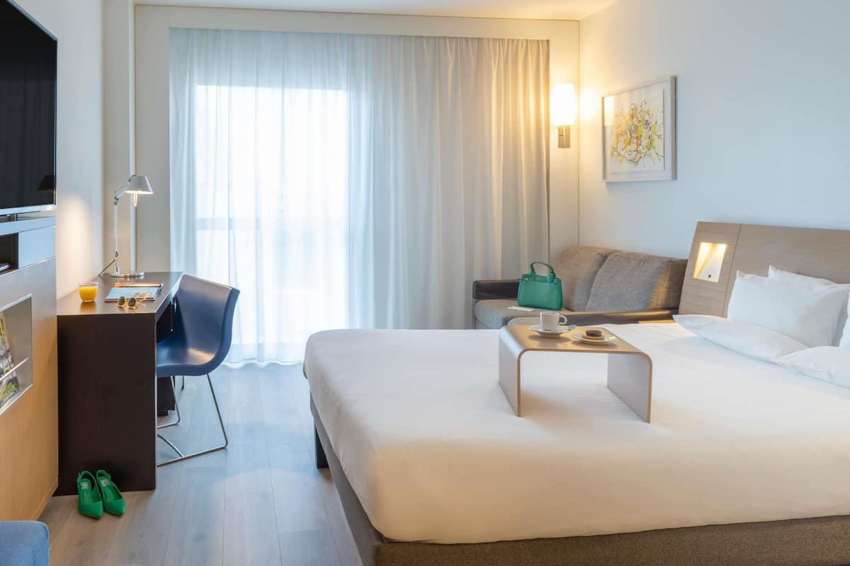 A hotel room with a bed, a desk with a chair, a television, and a window with curtains.