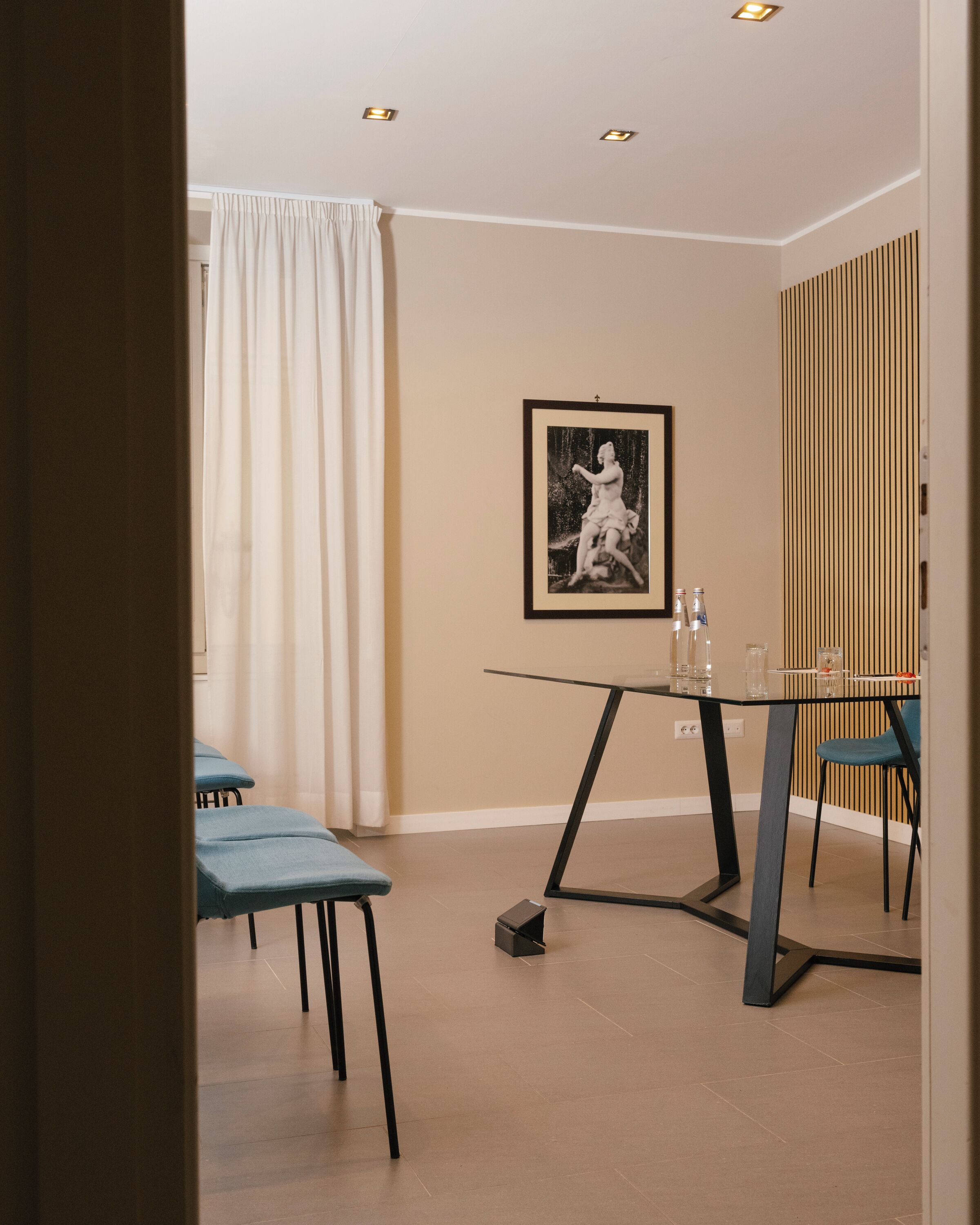 Una habitación moderna con una mesa de vidrio, sillas azules, un cuadro en la pared y un panel de madera en un lateral.