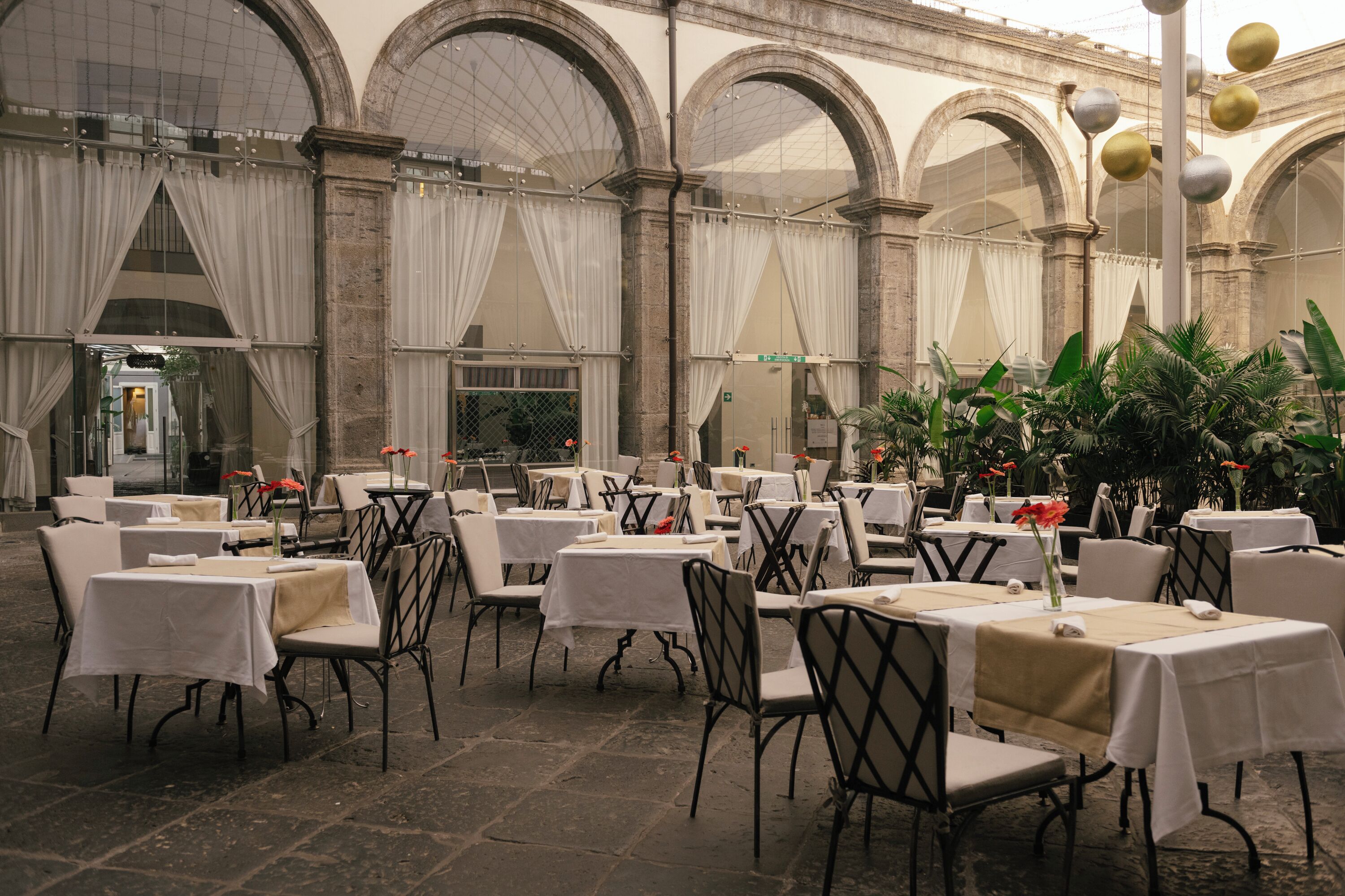An indoor dining area with tables and chairs, large windows with drapes, and a stone floor.