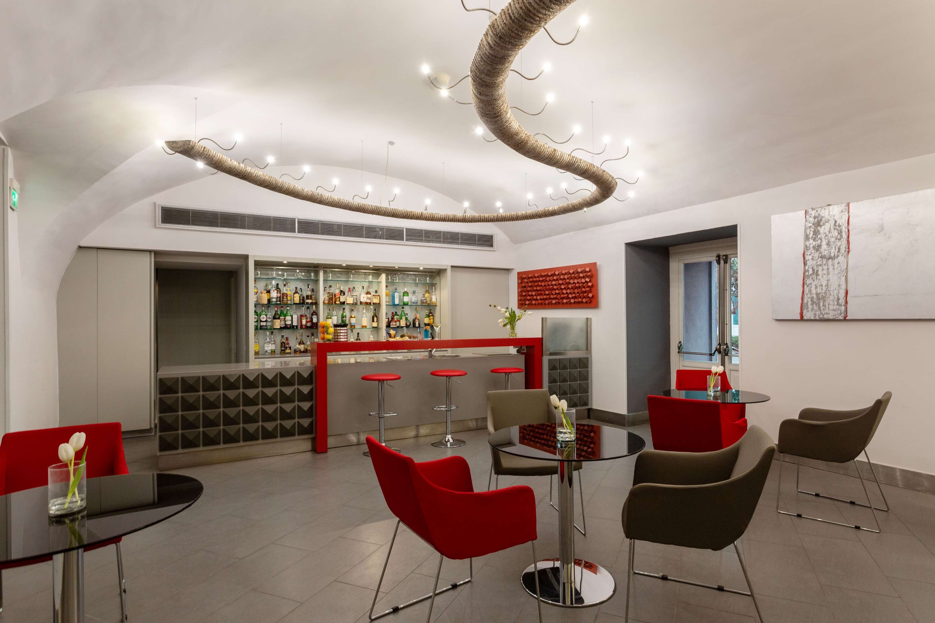 A modern bar area with a curved ceiling light fixture, bar stools, and a variety of bottles on the shelves.
