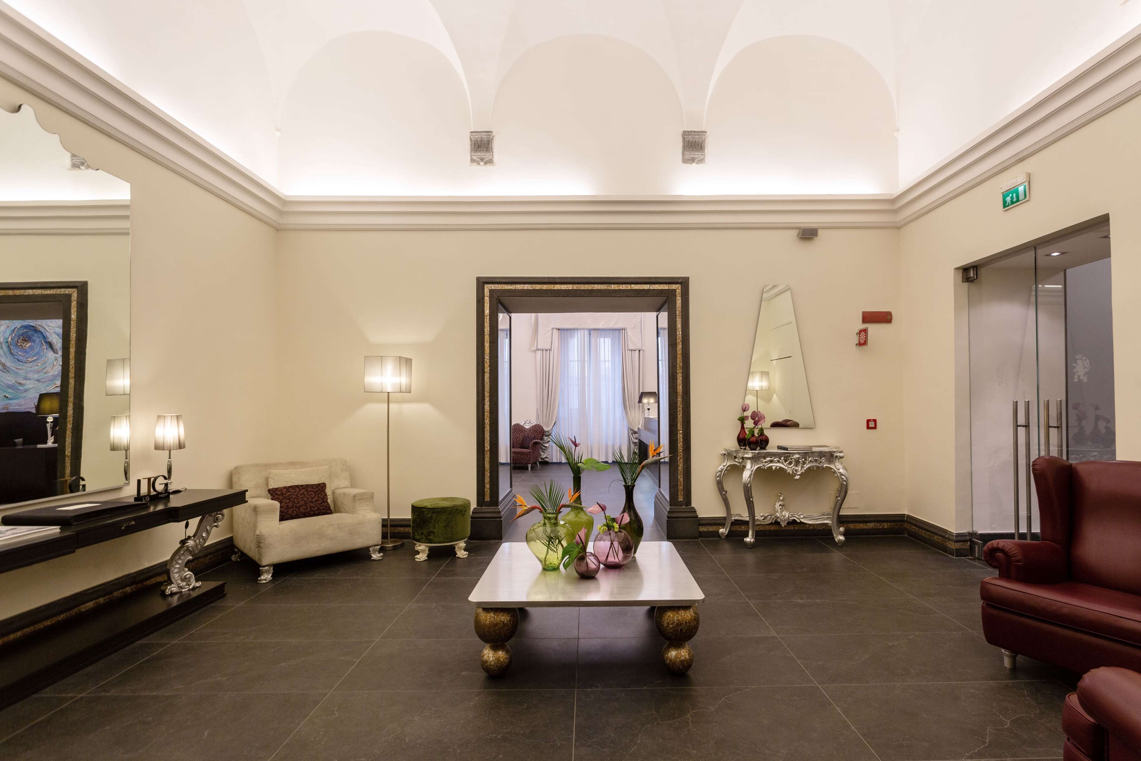 Una hall con una reception, un divano bianco, un divano rosso, un tavolino con vasi e uno specchio.