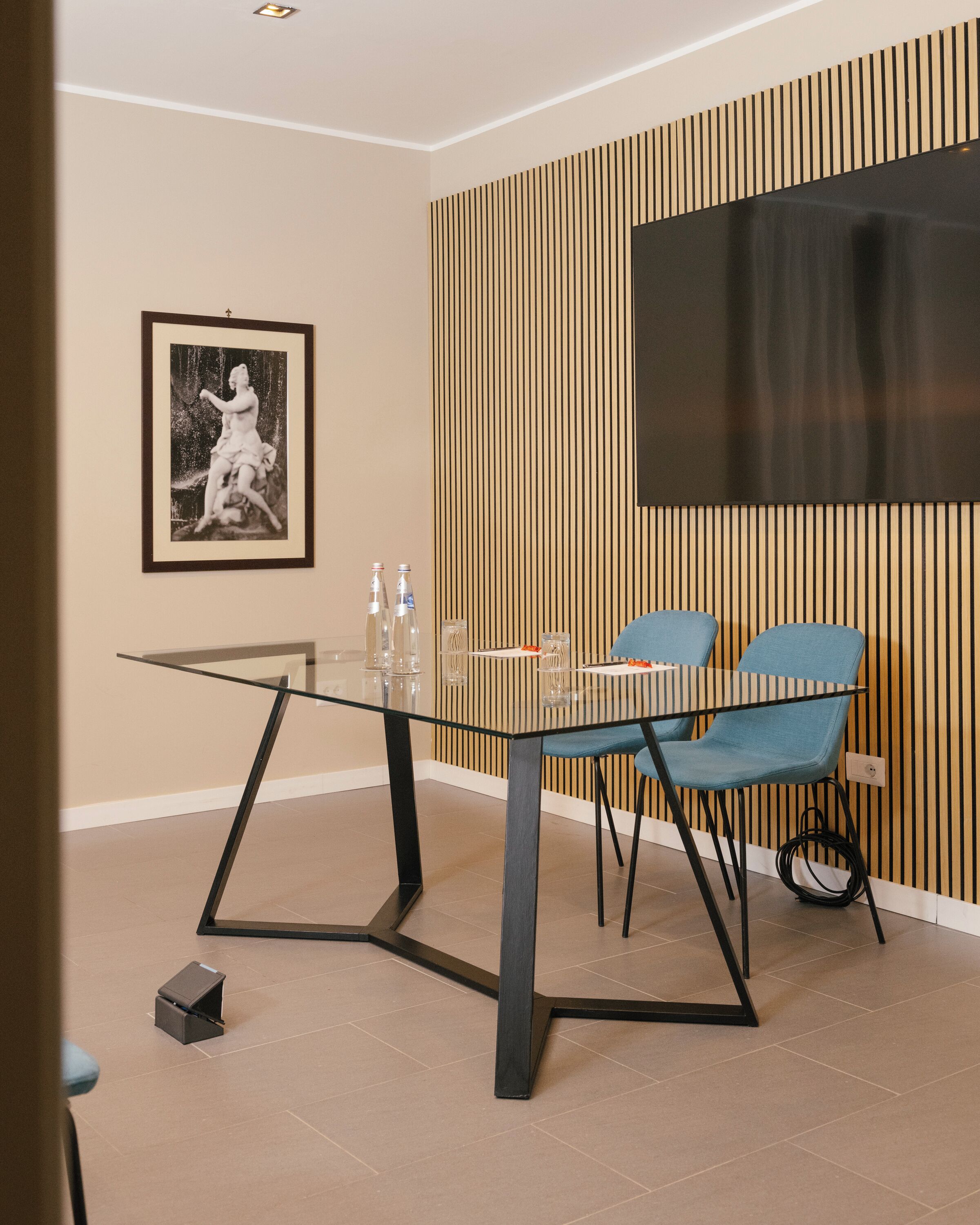 A modern dining area with a glass table, two blue chairs, a framed picture on the wall, and a wooden panel design.