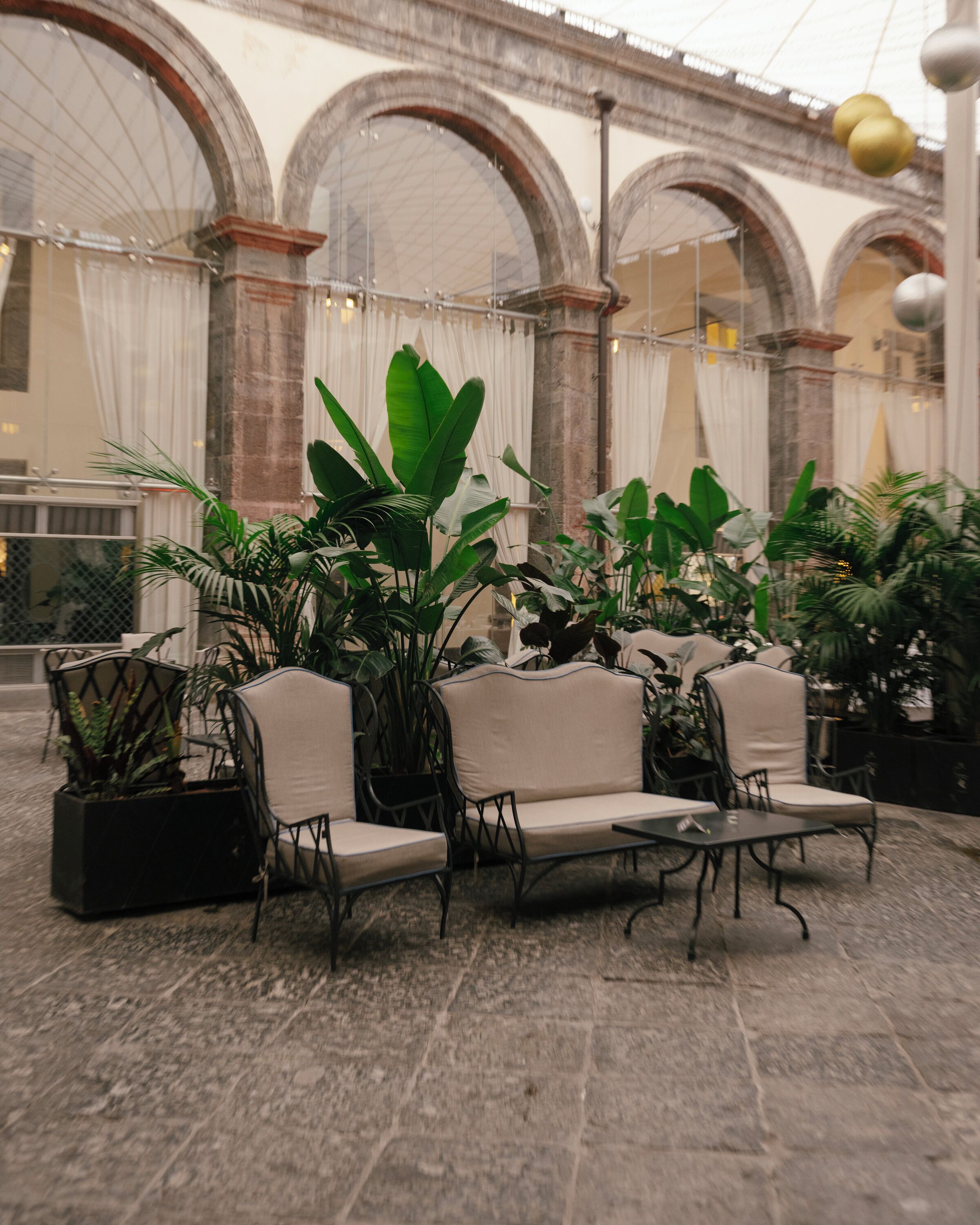 Un área de estar con sillas y mesas, rodeada de plantas en macetas, en un edificio con ventanas arqueadas y suelo de piedra.
