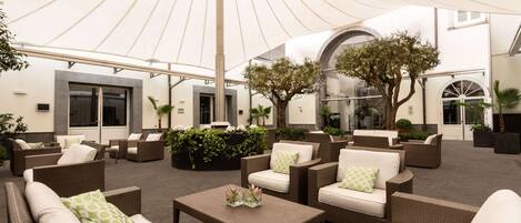 An outdoor seating area with wicker furniture and a large umbrella, surrounded by plants and a building with arched windows.